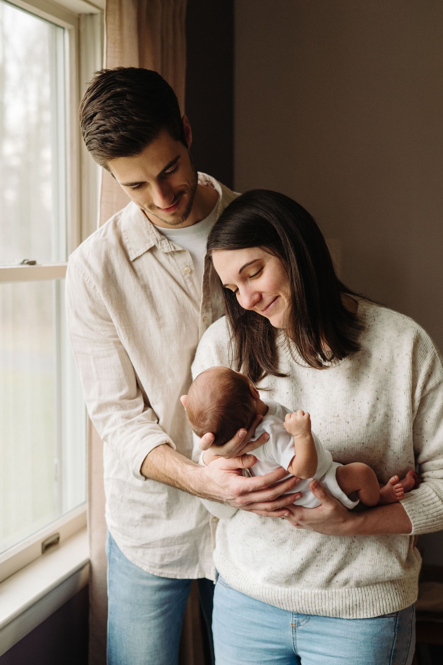 Young couple smiling and holding their newborn baby by a bright window in a cozy home setting.