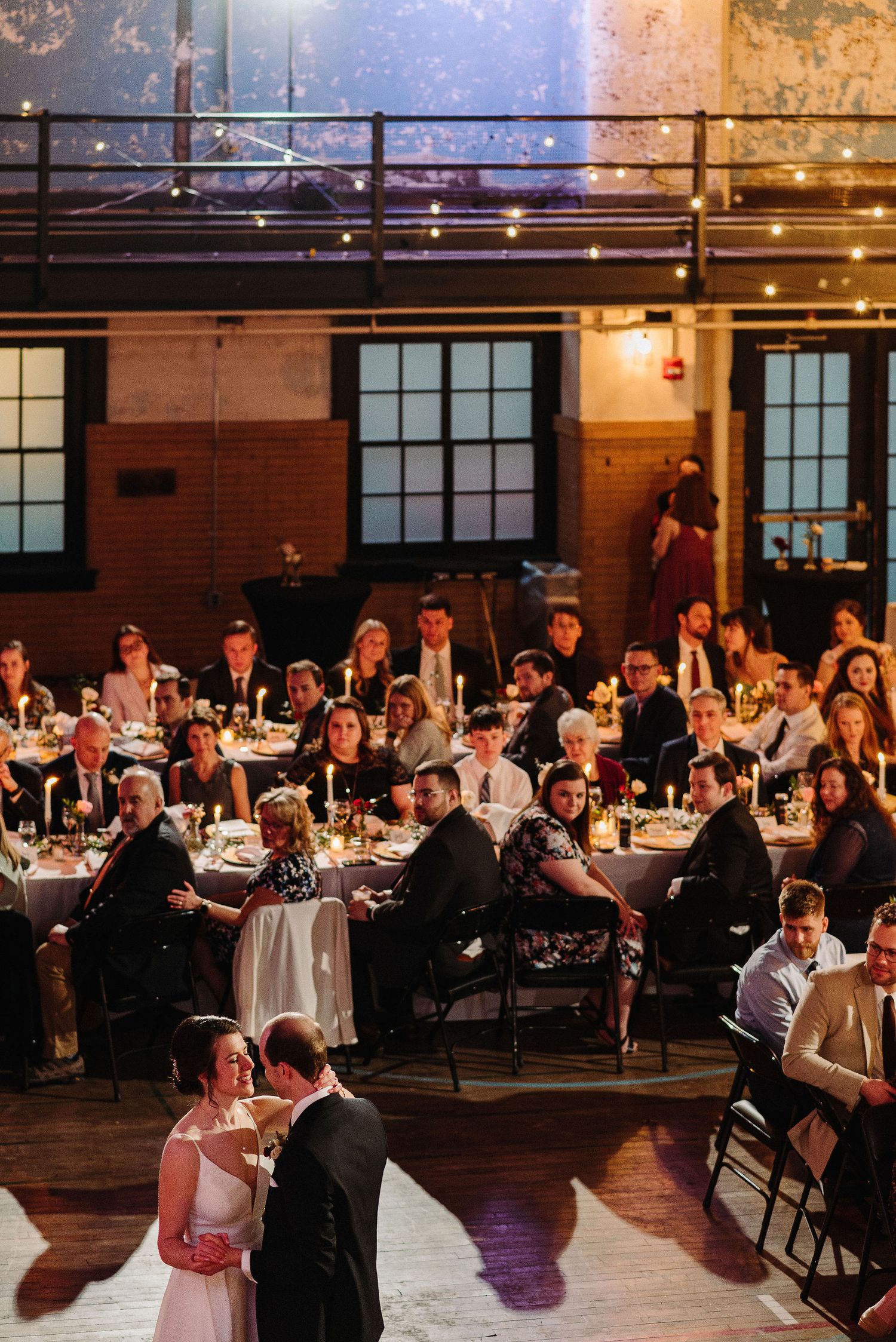 Wedding reception guests seated at long tables in an industrial-style venue with warm lighting and rustic decor.