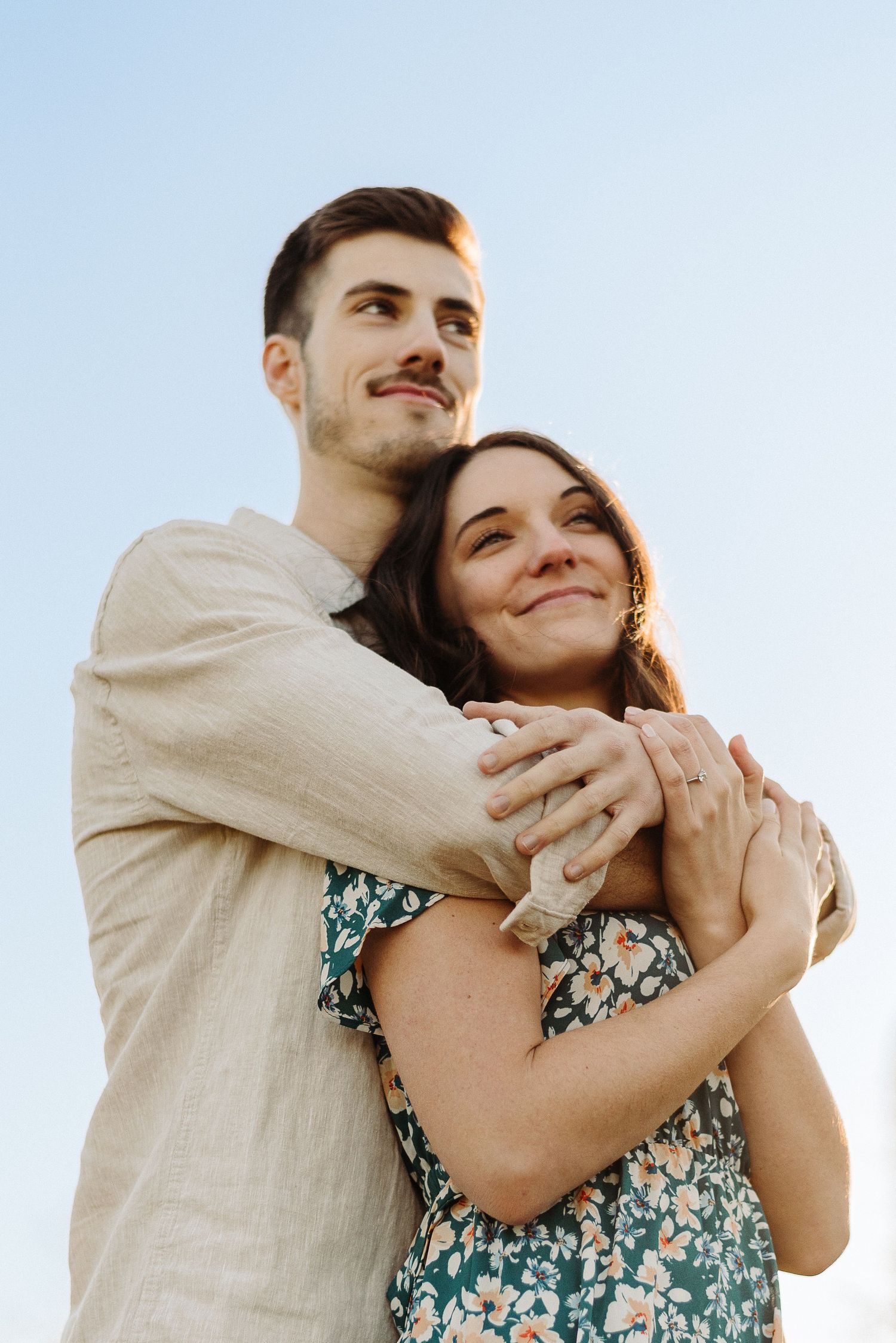 A happy couple embracing outdoors, man hugging woman from behind, both smiling upward against a clear blue sky.
