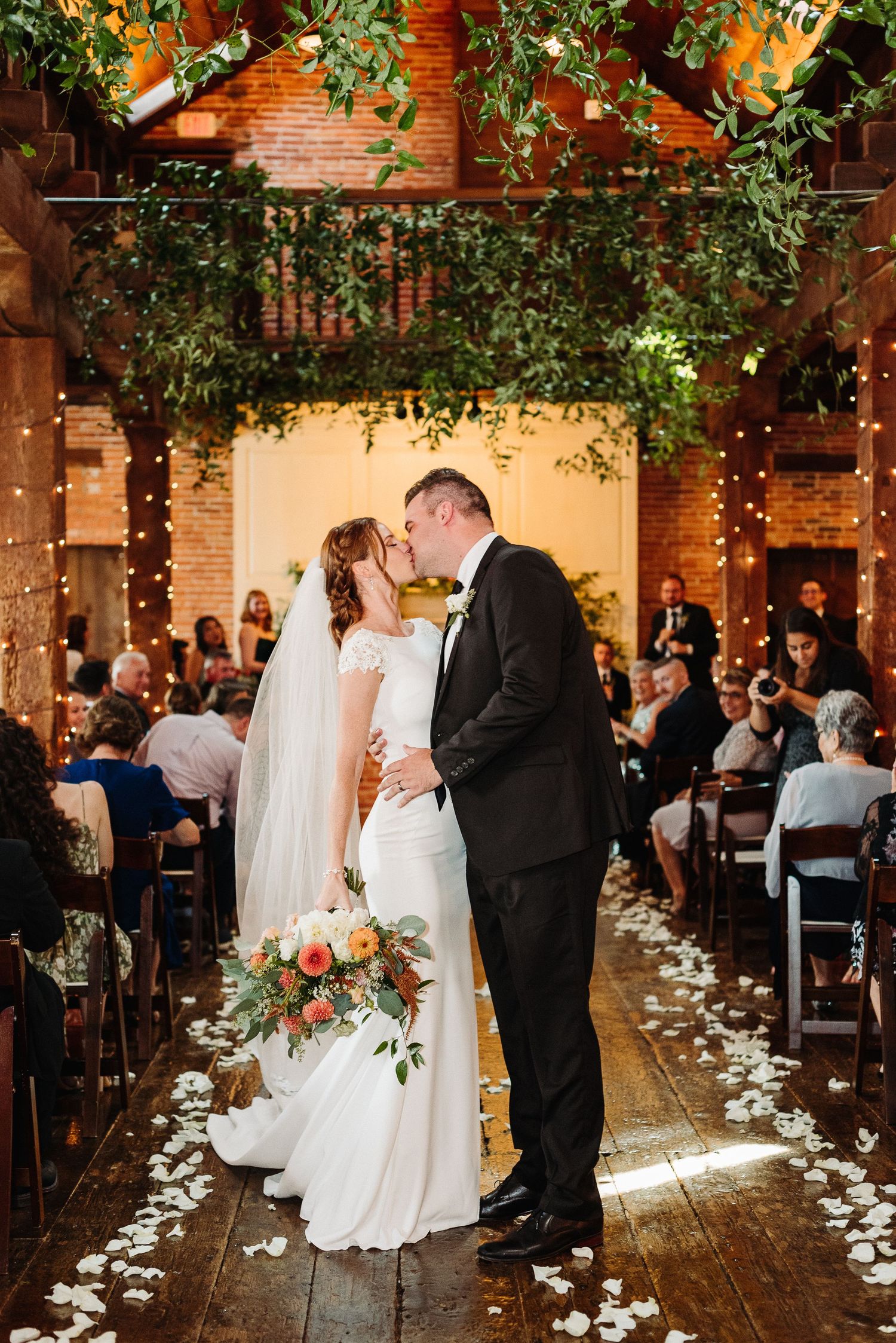 Wedding ceremony in rustic indoor venue with exposed brick walls and scattered white rose petals along wooden aisle in Manheim PA.