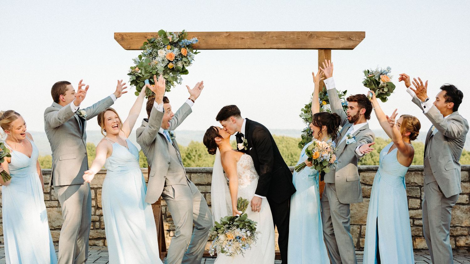 Wedding party in gray suits and light blue dresses pose together in a joyful celebration lineup outdoors.
