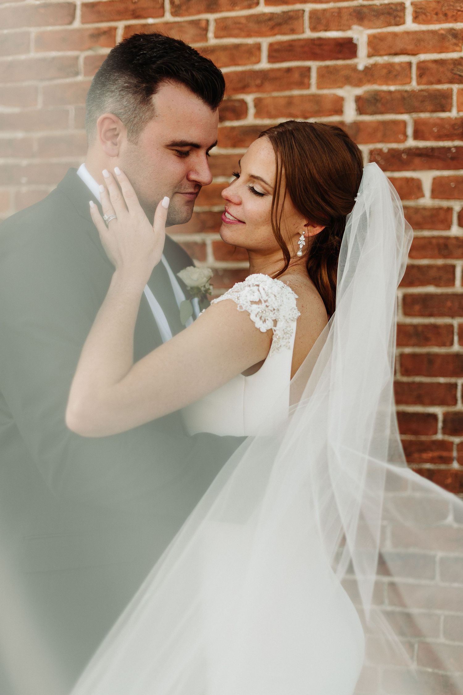 A romantic wedding couple embraces against a rustic brick wall backdrop with soft lighting and flowing white veil.