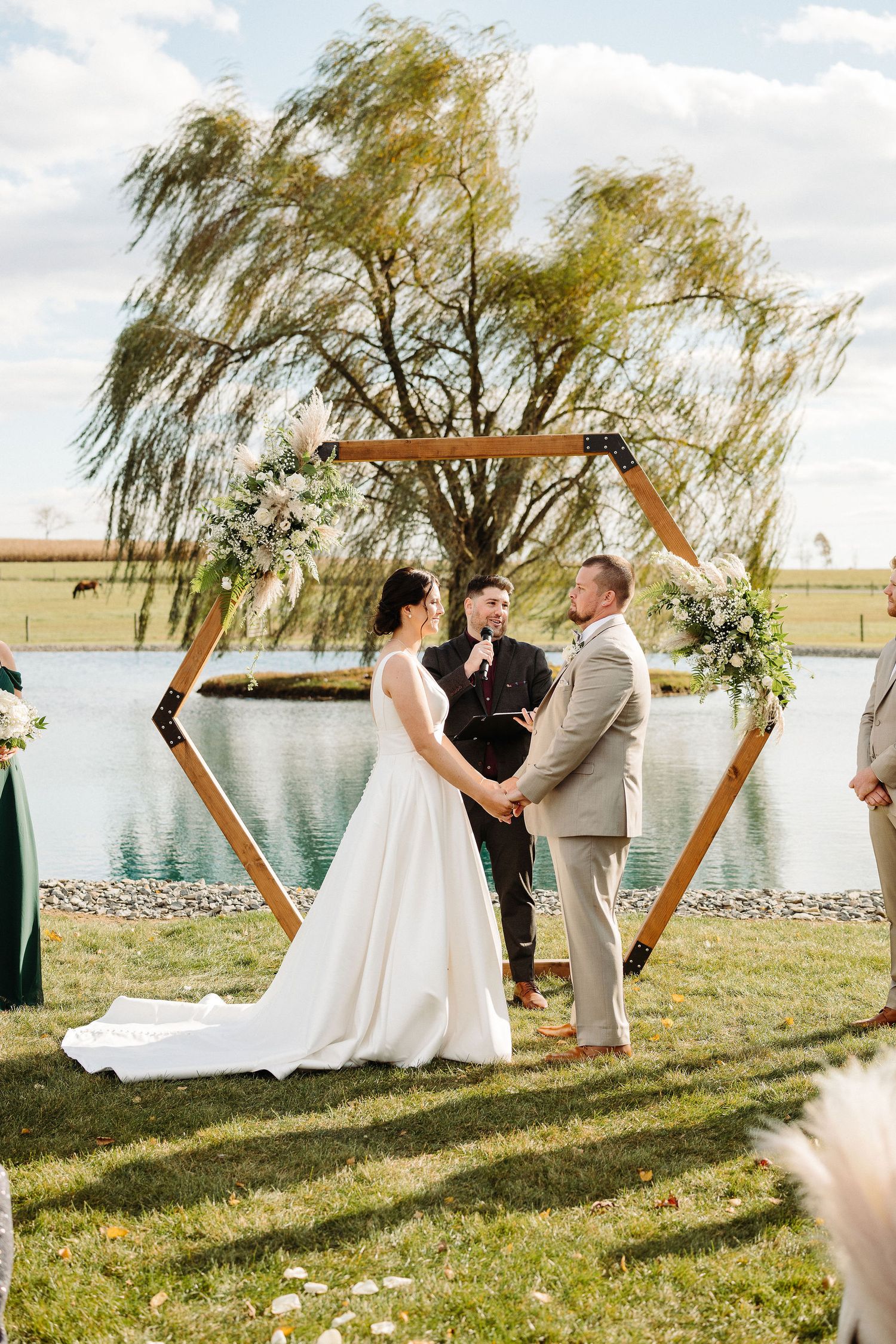 Wedding ceremony takes place by a tranquil lake with willow trees and a wooden hexagonal arch at sunset.