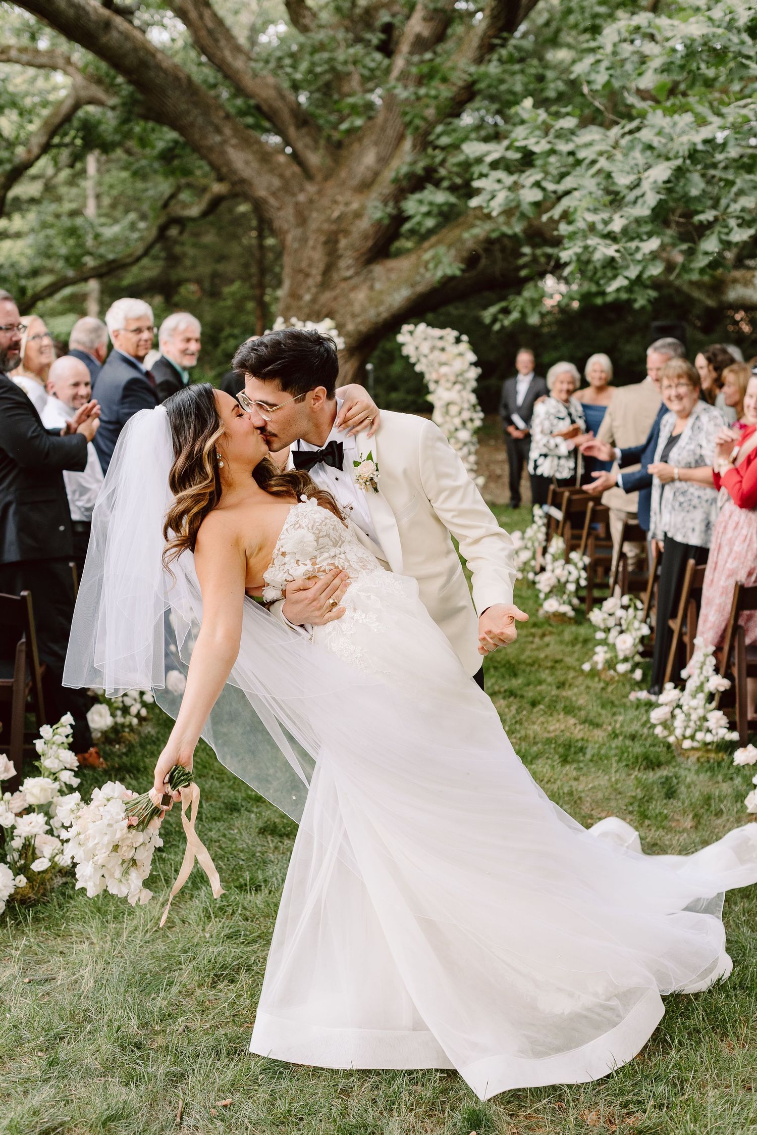 Bride and groom share a romantic dip kiss at outdoor wedding ceremony with floral aisle and guests applauding.