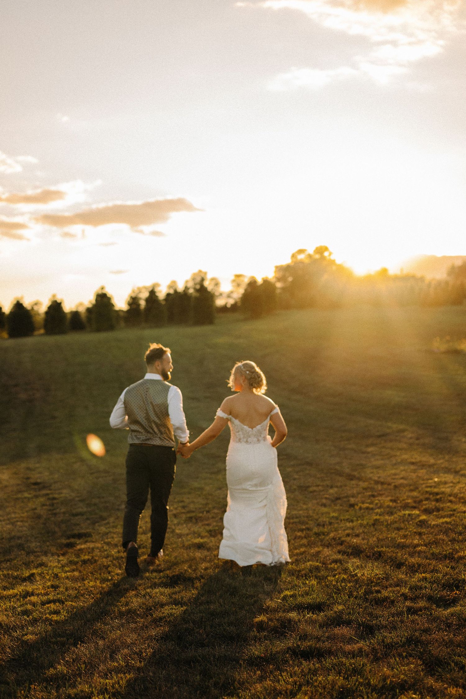 A couple walks hand in hand across a golden field at sunset, with sunbeams and silhouetted trees in the background.