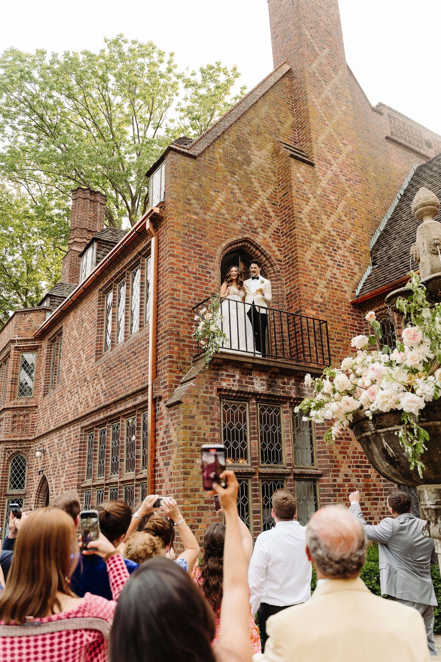 A crowd gathers outside brick buildings to watch someone toss a wedding bouquet in a celebratory moment.