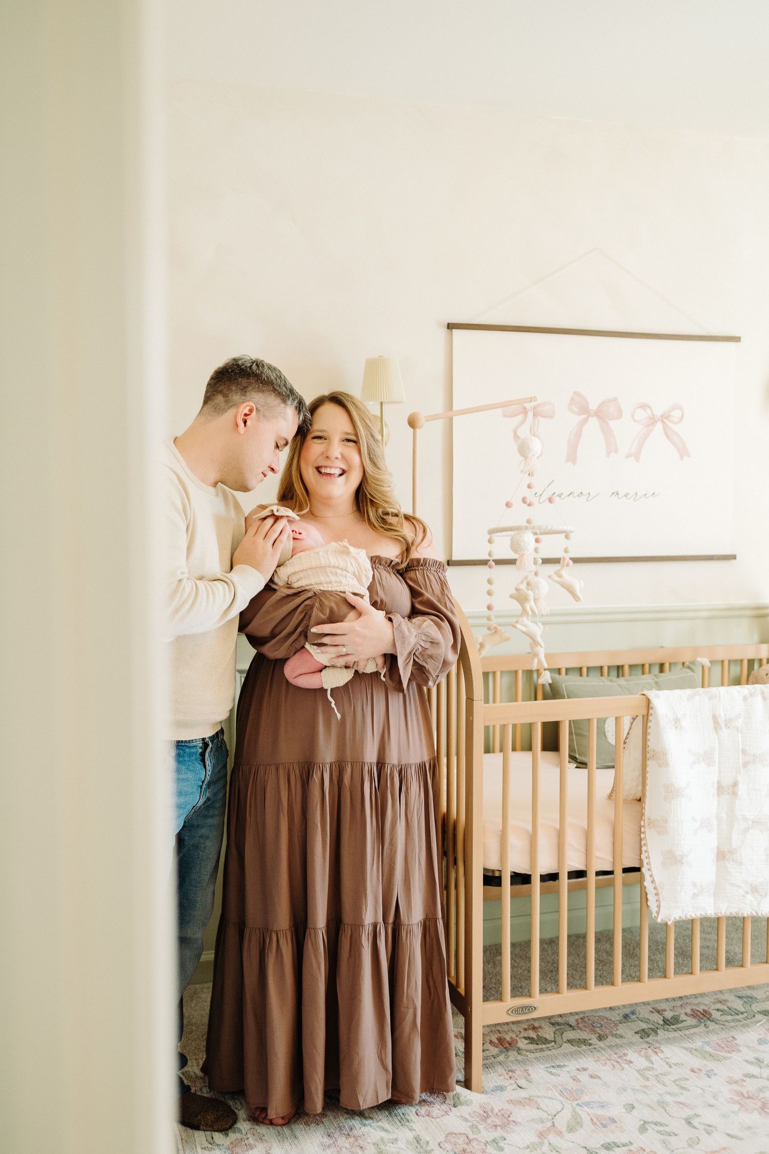 A couple stands lovingly in a nursery with a wooden crib and pink wall art, sharing a tender moment together.