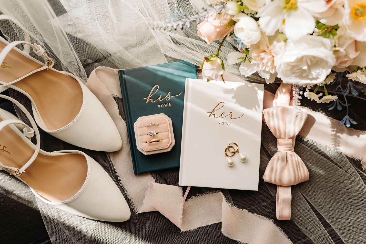Wedding accessories including white heels, invitations, rings, and delicate pink bows arranged on a dark surface.