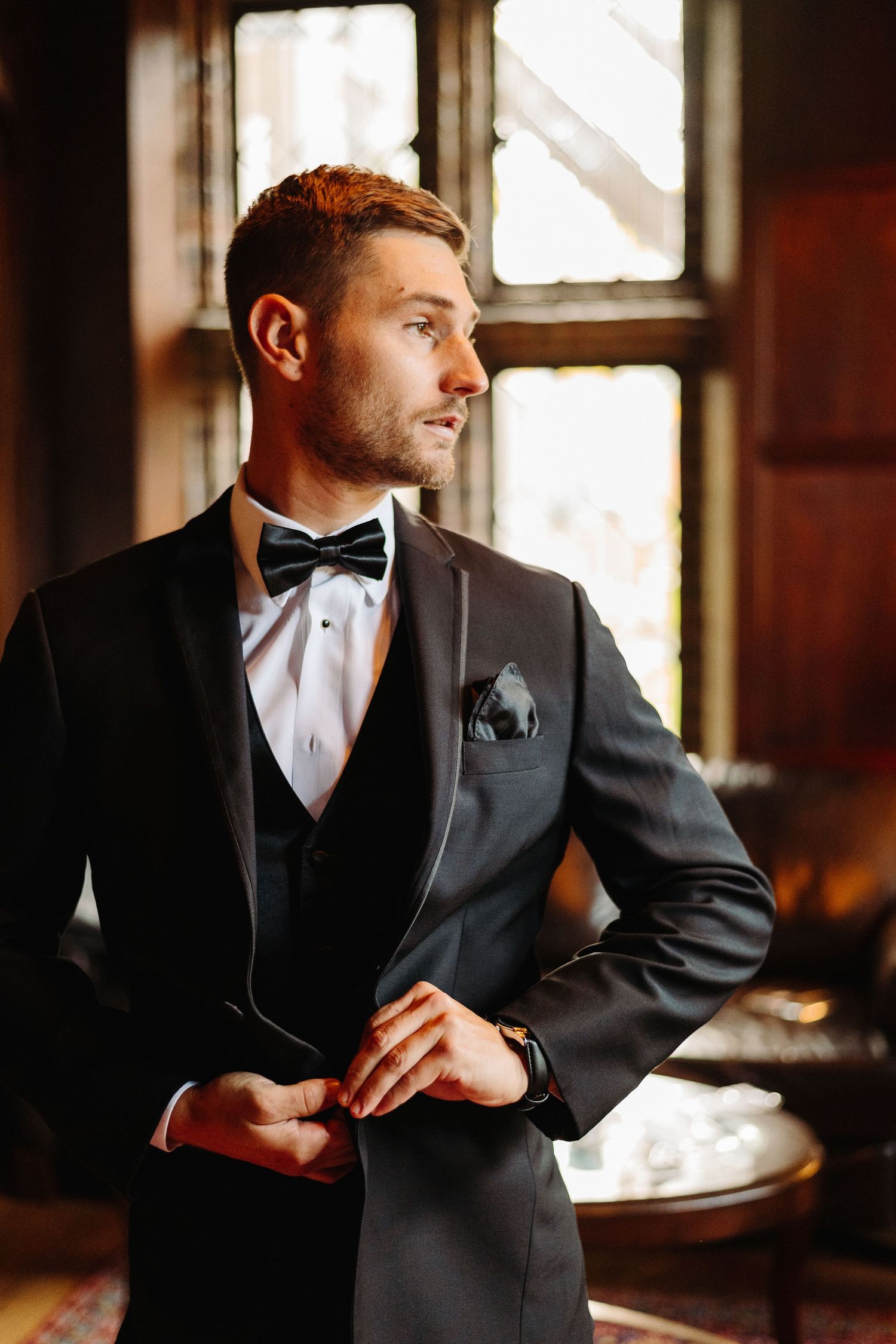 A groom in a black tuxedo and bow tie poses in an elegant indoor setting with natural window lighting.