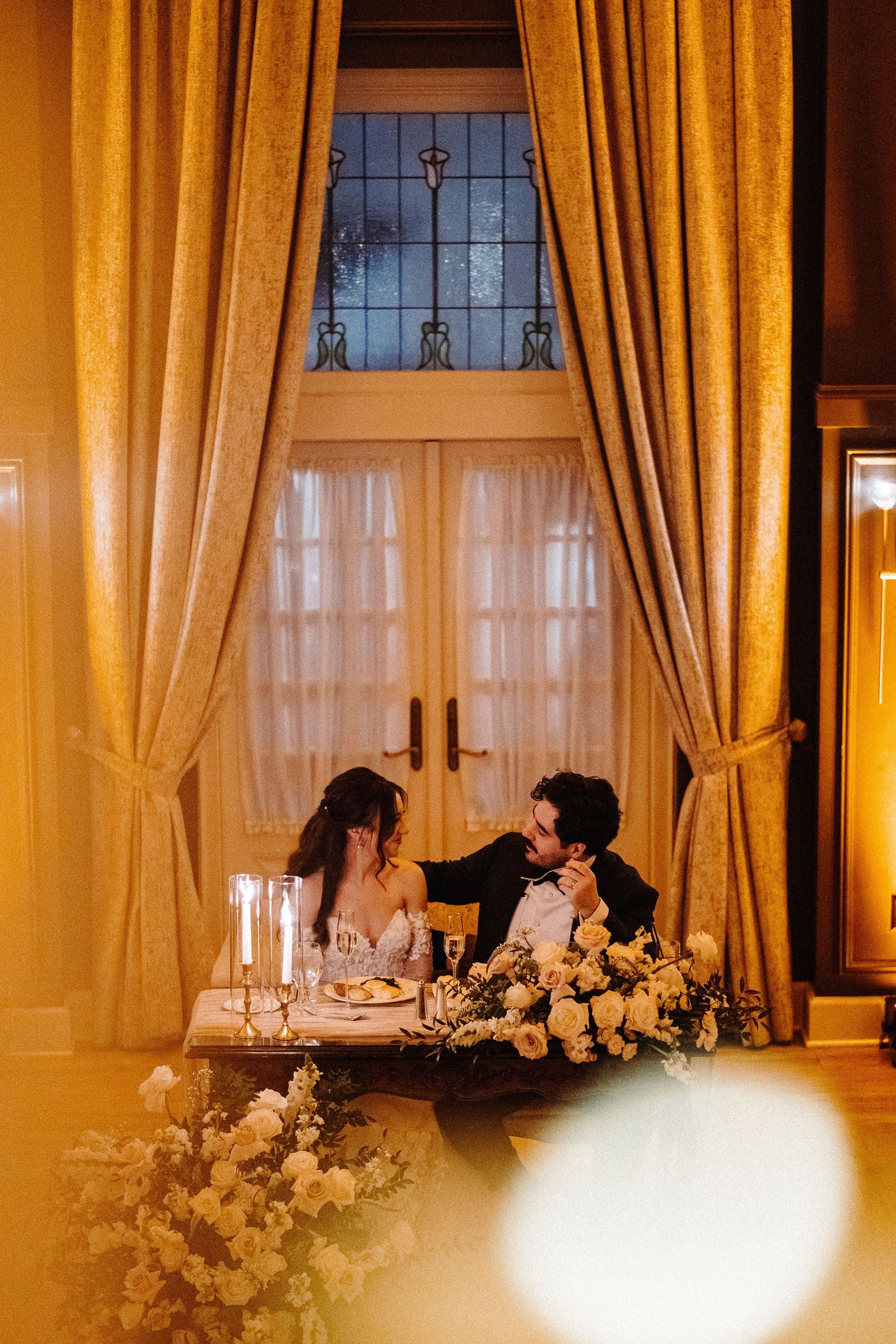 Elegant wedding reception table setting with warm lighting, golden curtains, and white floral centerpieces.
