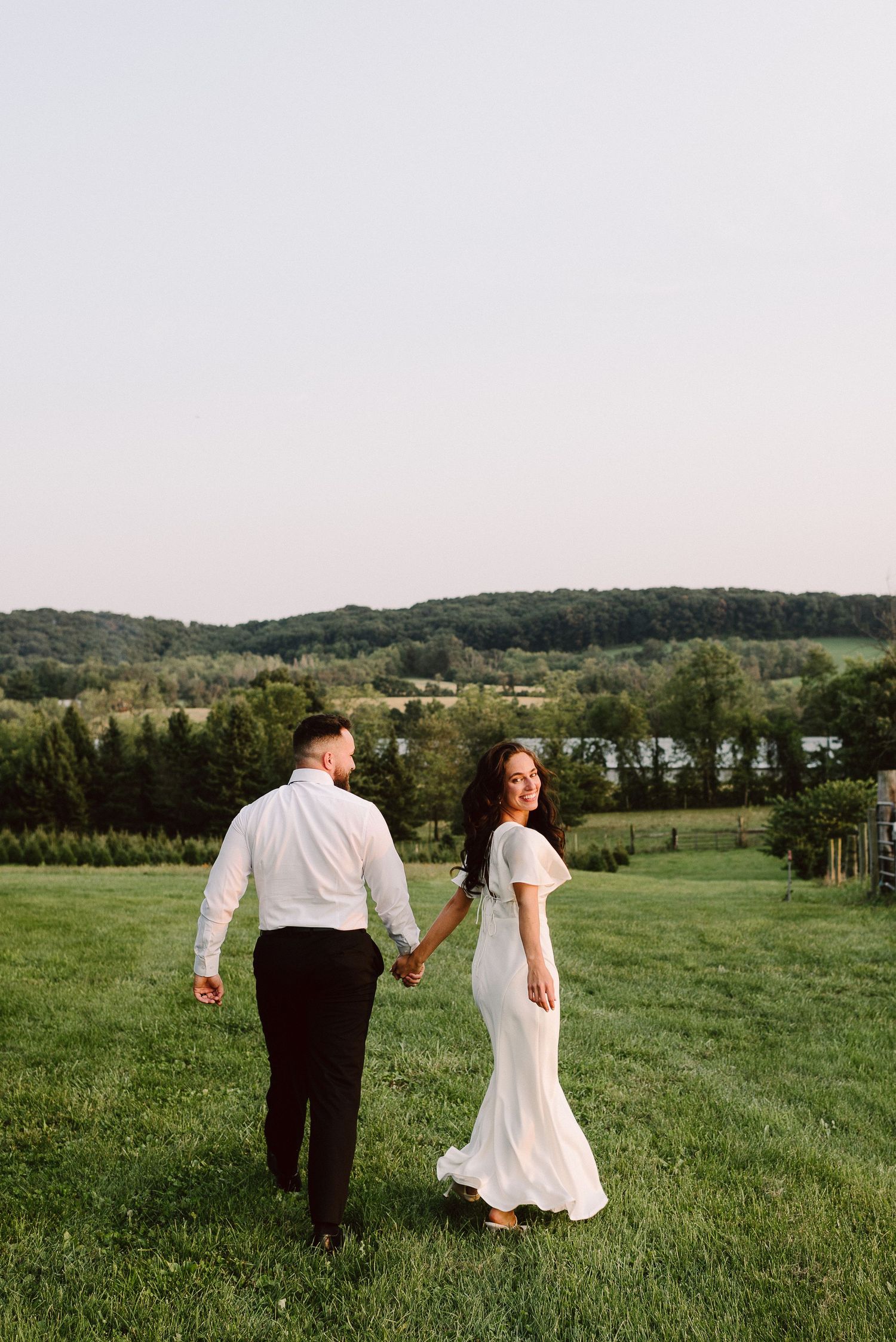 A couple walks hand in hand through a lush green field with rolling hills and trees in the background at sunset.
