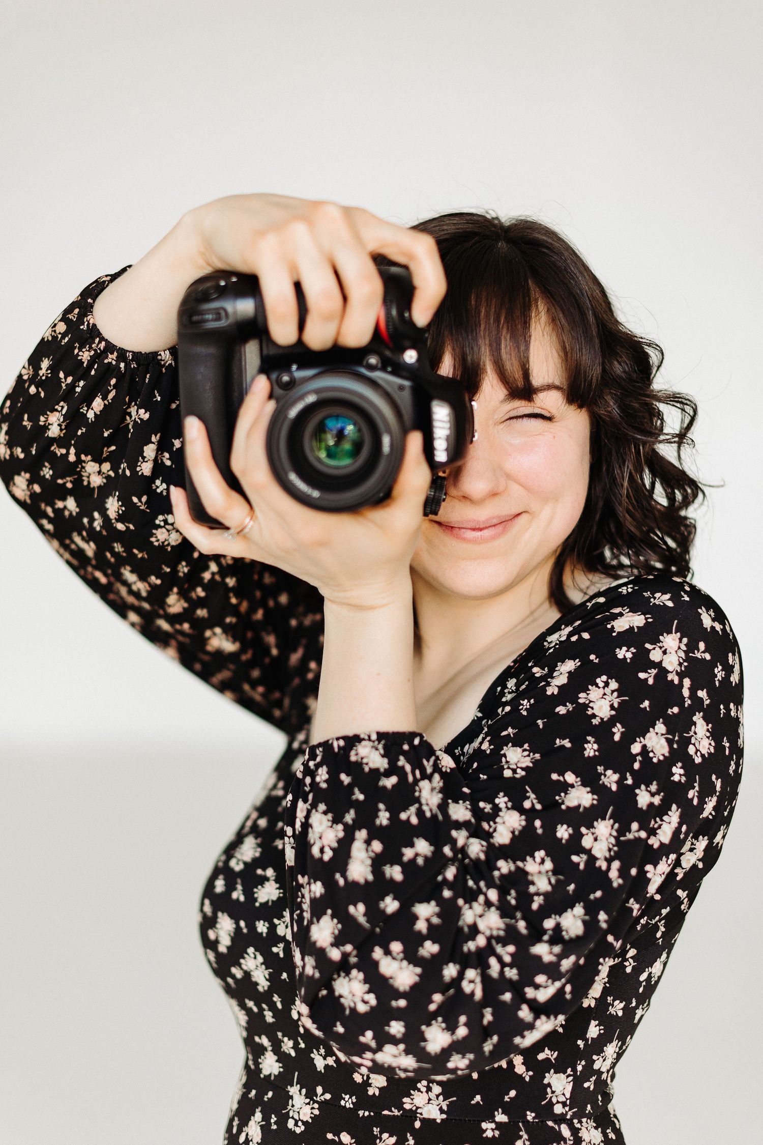 Photographer in a floral patterned dress uses professional DSLR camera against white background.