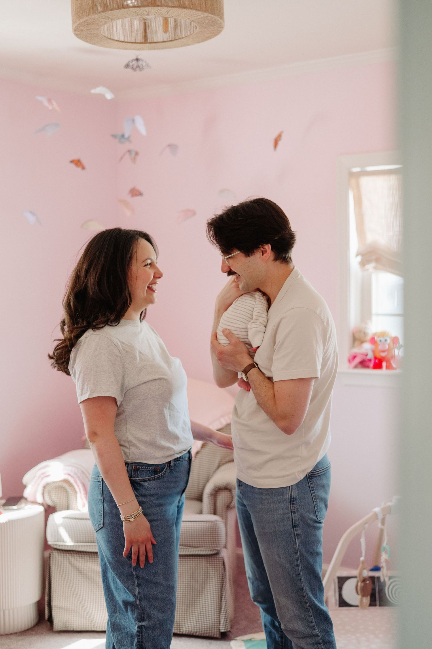 A couple in matching white tees and jeans share a tender moment holding their newborn in a pink nursery.