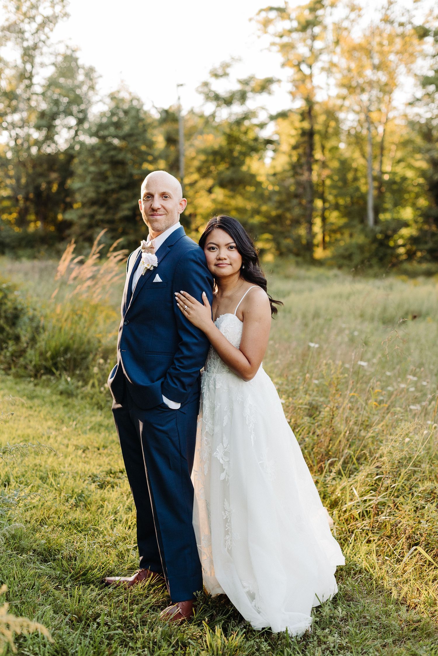 A couple poses together on their wedding day in a sunny field with glowing autumn trees in the background.