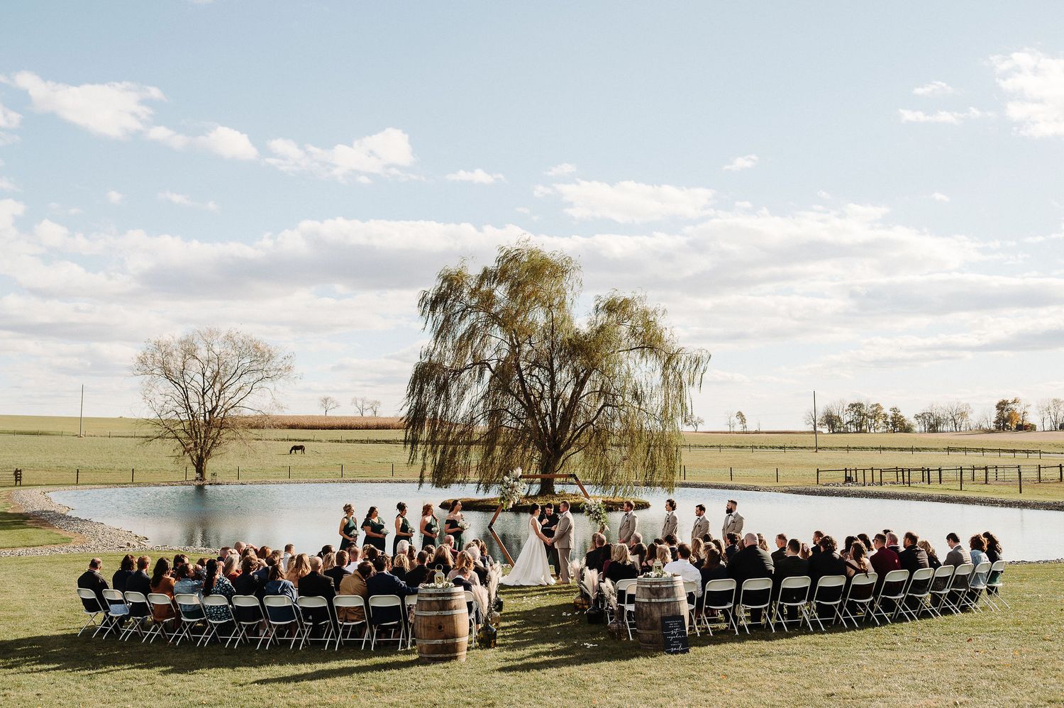 Wedding party sits in black chairs arranged in a row outdoors with willow trees and a lake in the background.