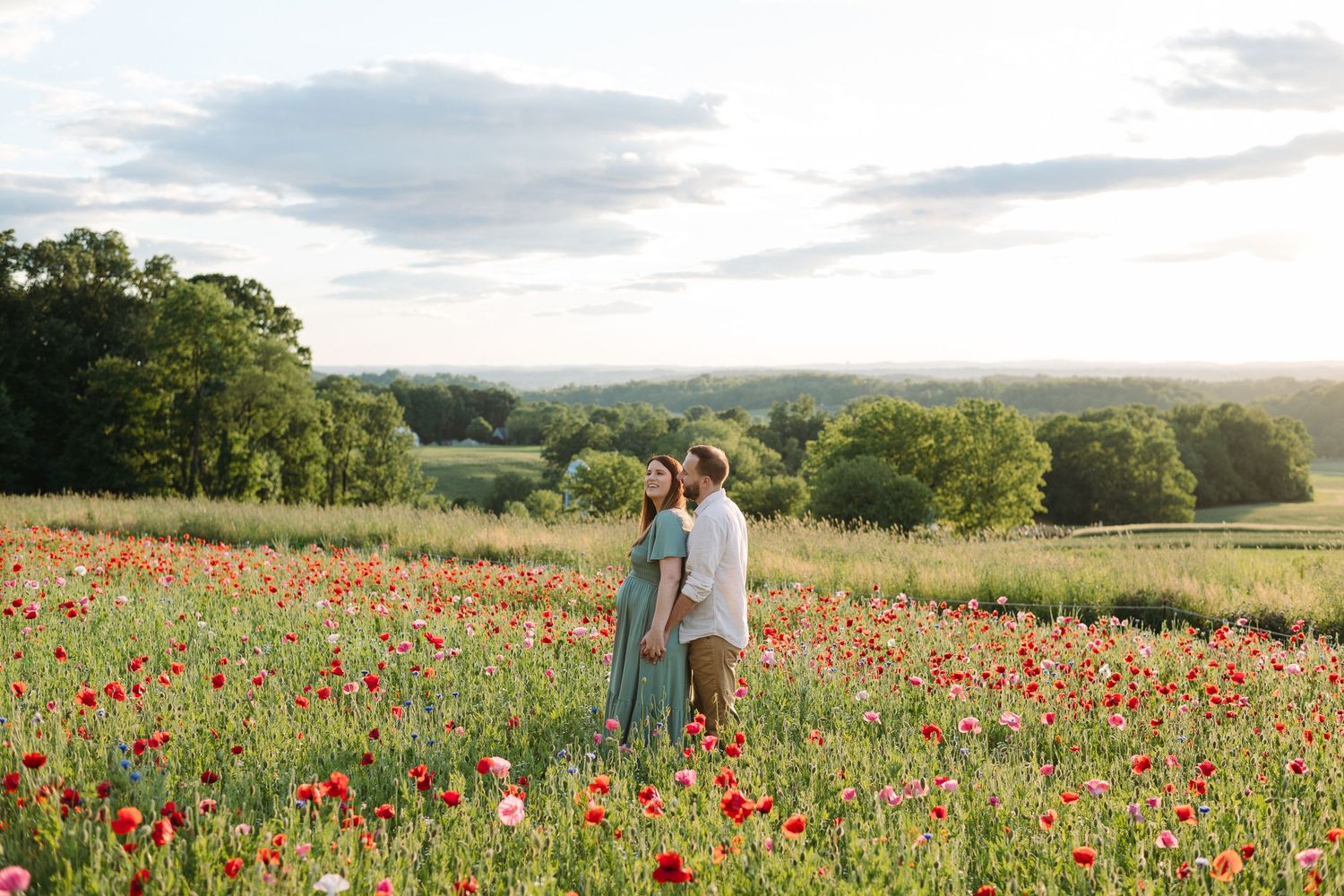 Couple standing in a vibrant red poppy field at golden hour with lush green trees and rolling hills in the background.