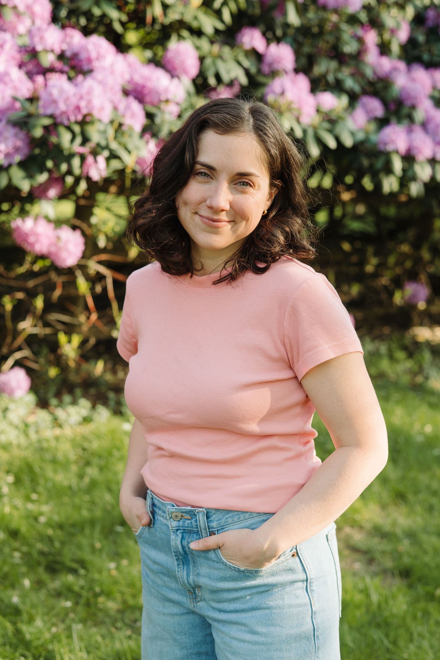 Young woman in pink t-shirt and jeans smiling outdoors near blooming pink rhododendron bushes in a garden.