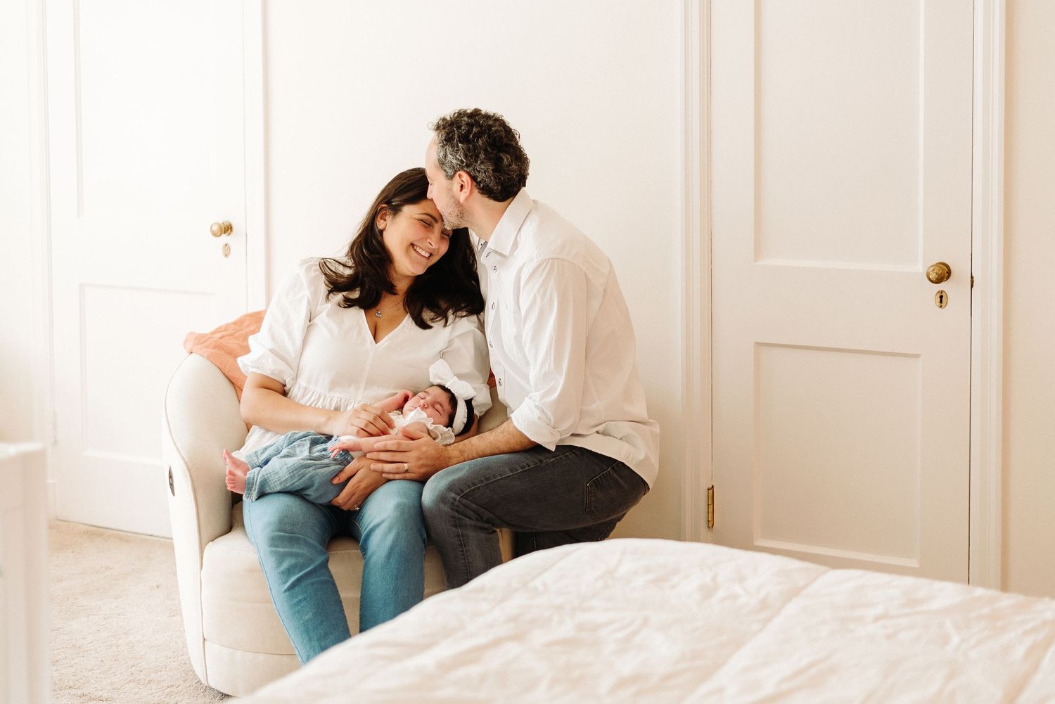A couple sits lovingly on a white armchair while holding their newborn baby in a bright, sunlit bedroom.