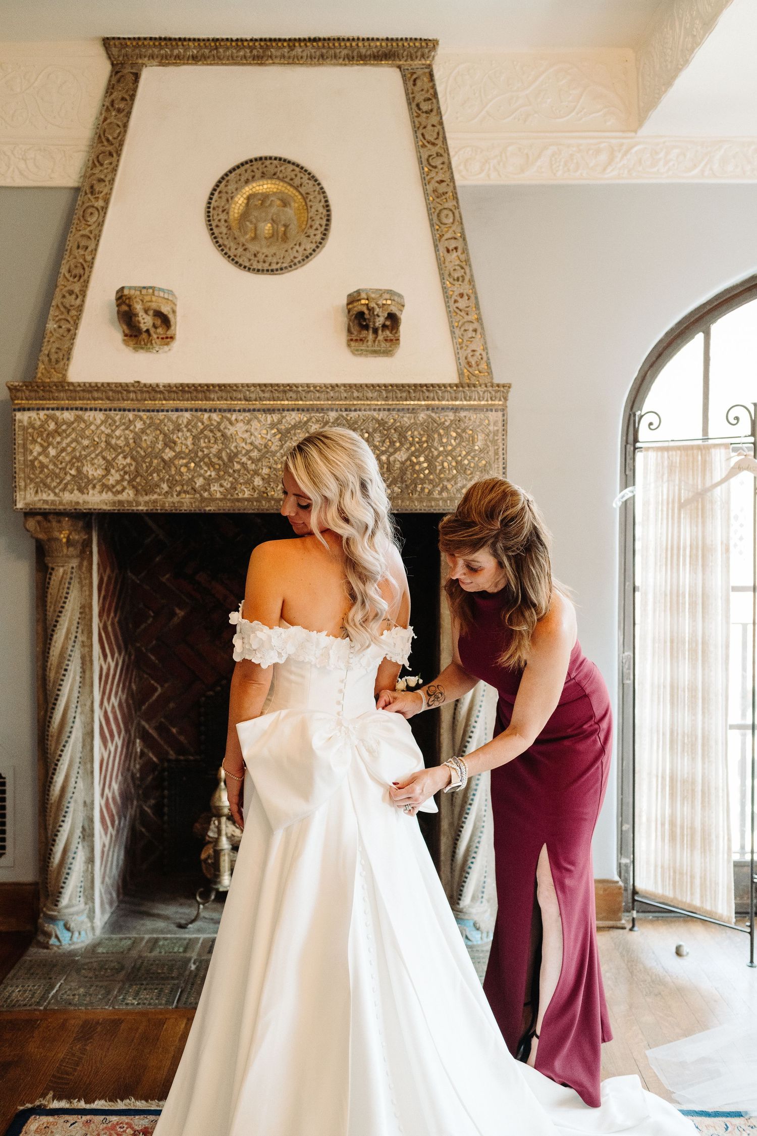 A wedding dress fitting moment captured in an elegant room with an ornate stone fireplace and arched doorway.