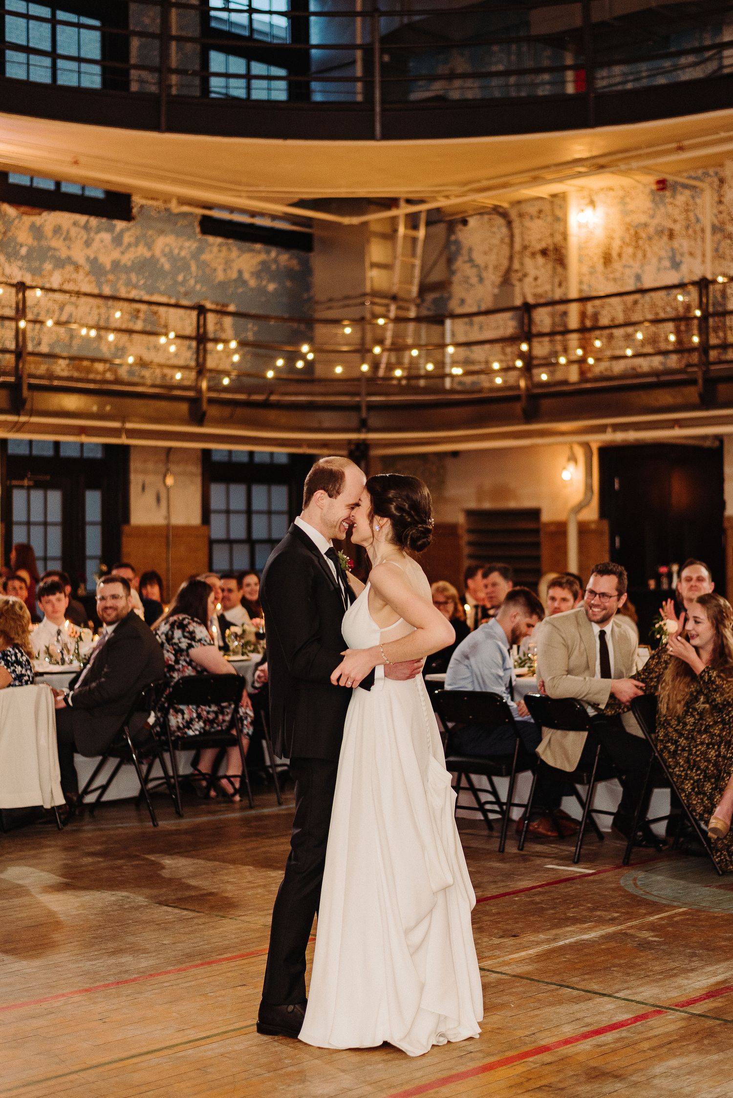 A couple shares their first dance at an industrial-style wedding venue with string lights and exposed brick walls.