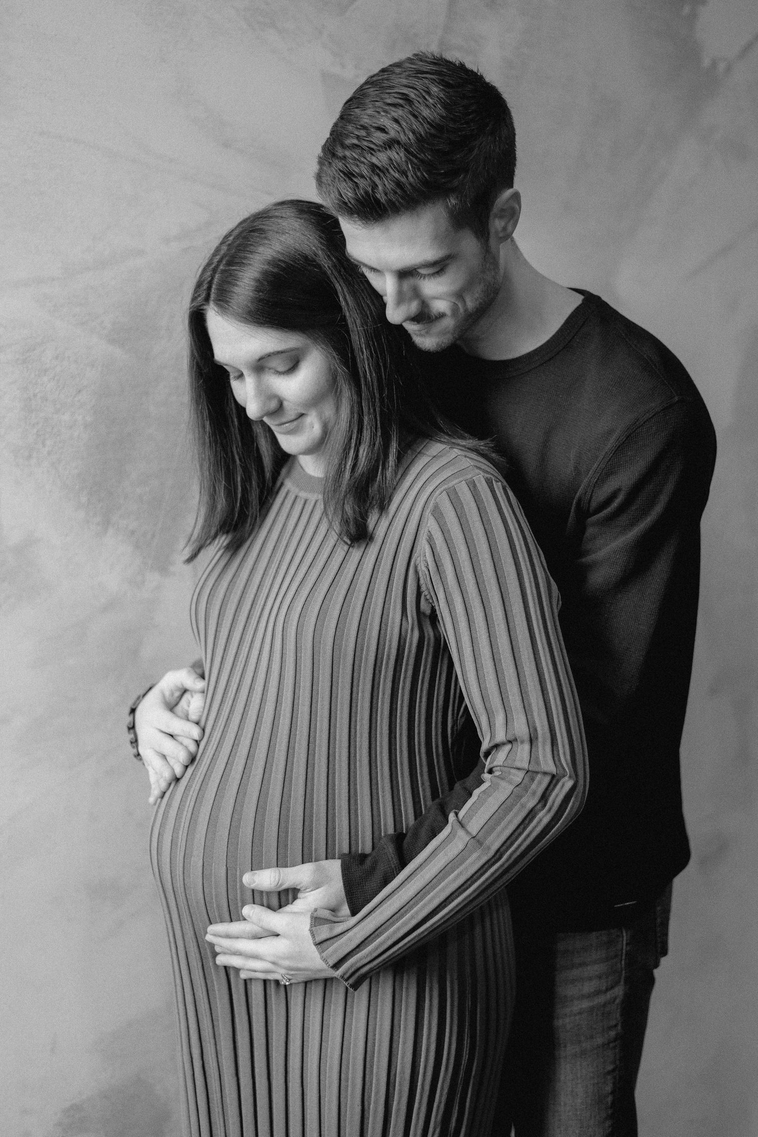 Black and white maternity photo of a couple tenderly cradling a pregnant belly in a studio setting.