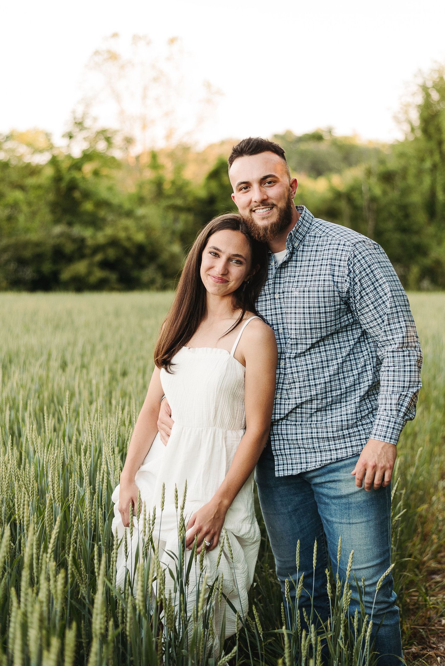 A couple stands together in a wheat field at sunset, wearing casual summer attire in Berks County PA.
