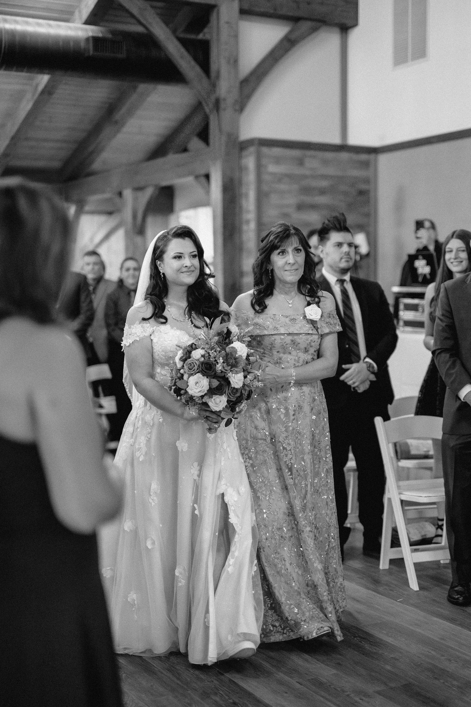Black and white wedding processional photo showing bridesmaids walking down the aisle in formal dresses at an indoor venue.