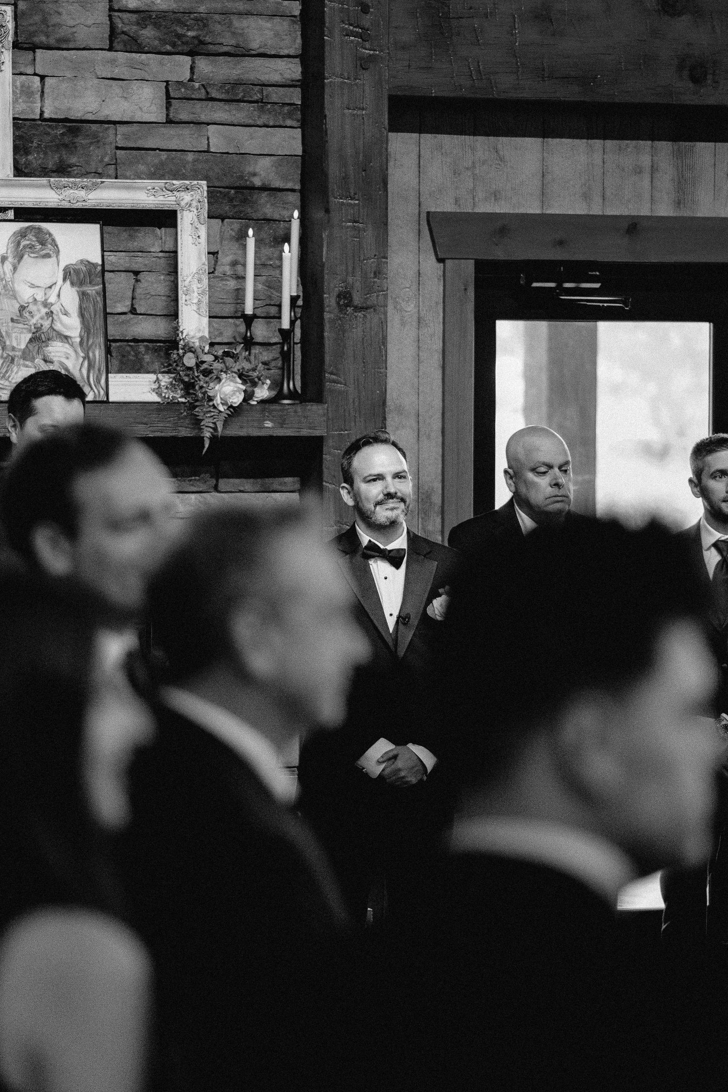 A black and white photograph shows a group of men in formal attire standing against a stone wall.