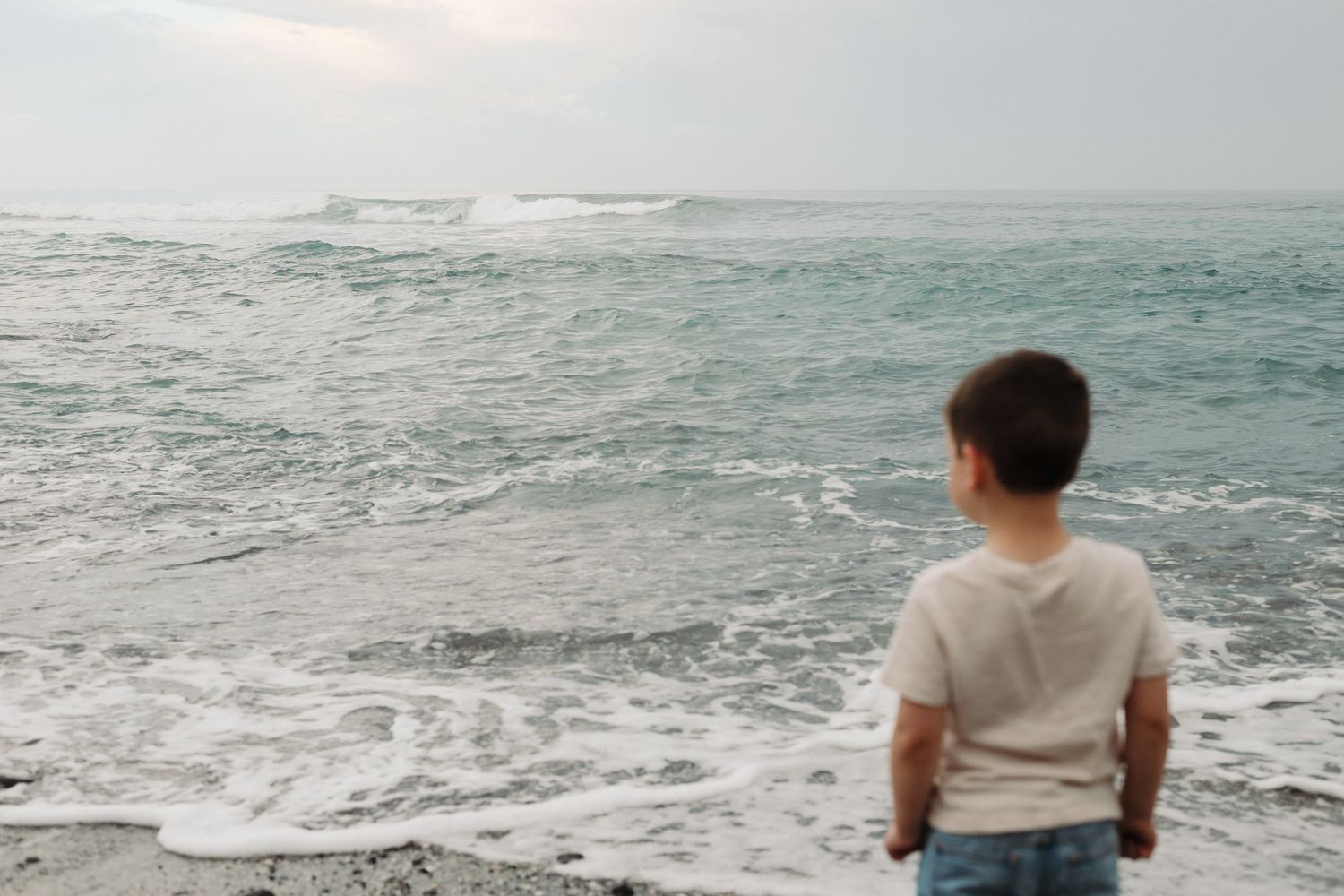 Person sitting on a rocky shore watching waves crash against the coastline on a cloudy day.