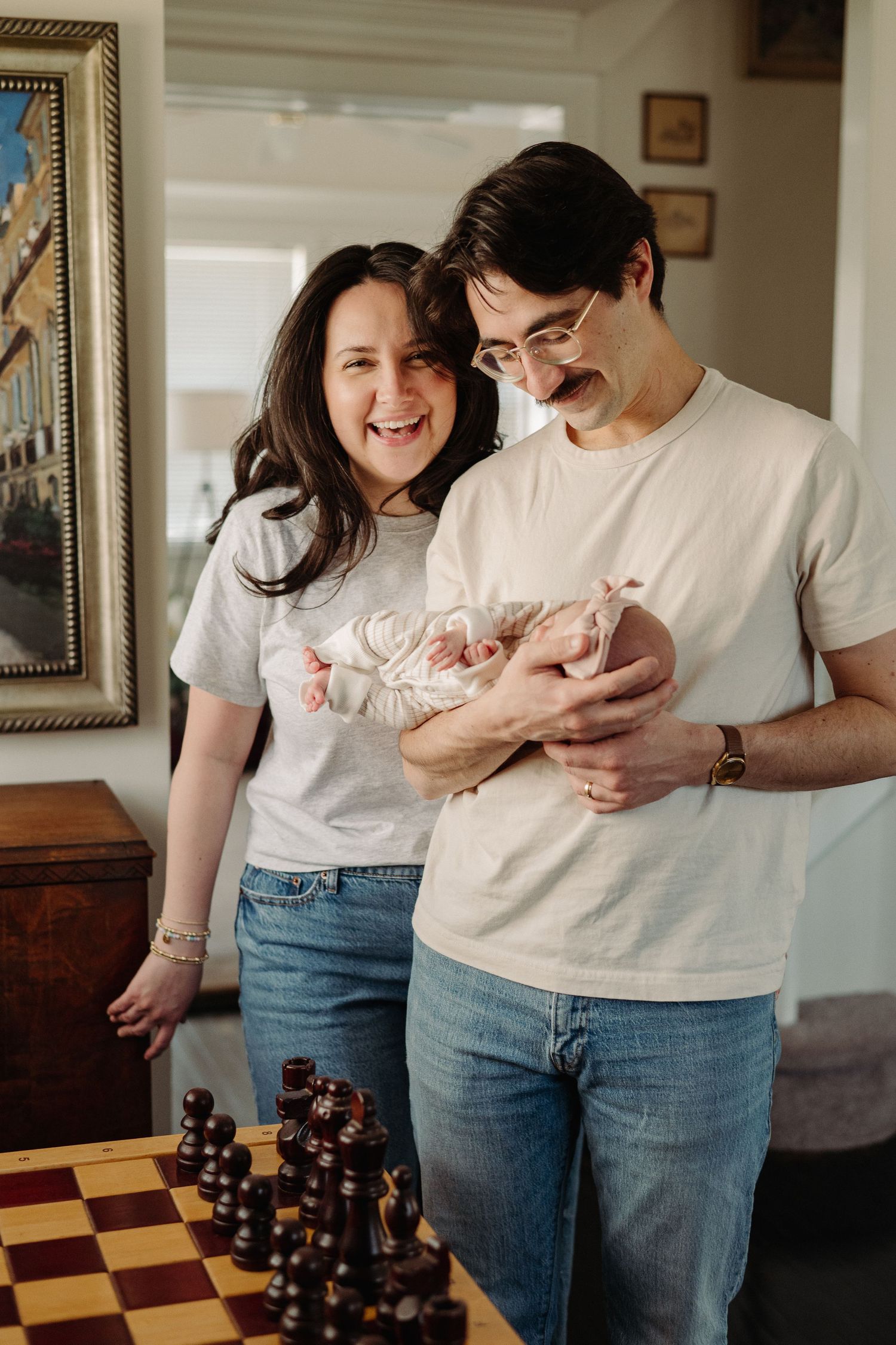 Happy couple in matching gray tees and jeans holding a newborn baby, standing near a chess board in a cozy home.