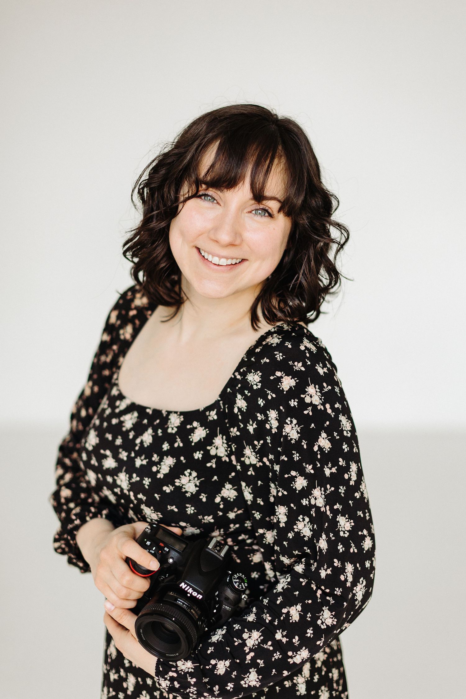 Professional photographer wearing floral print dress stands with Nikon camera against white background.