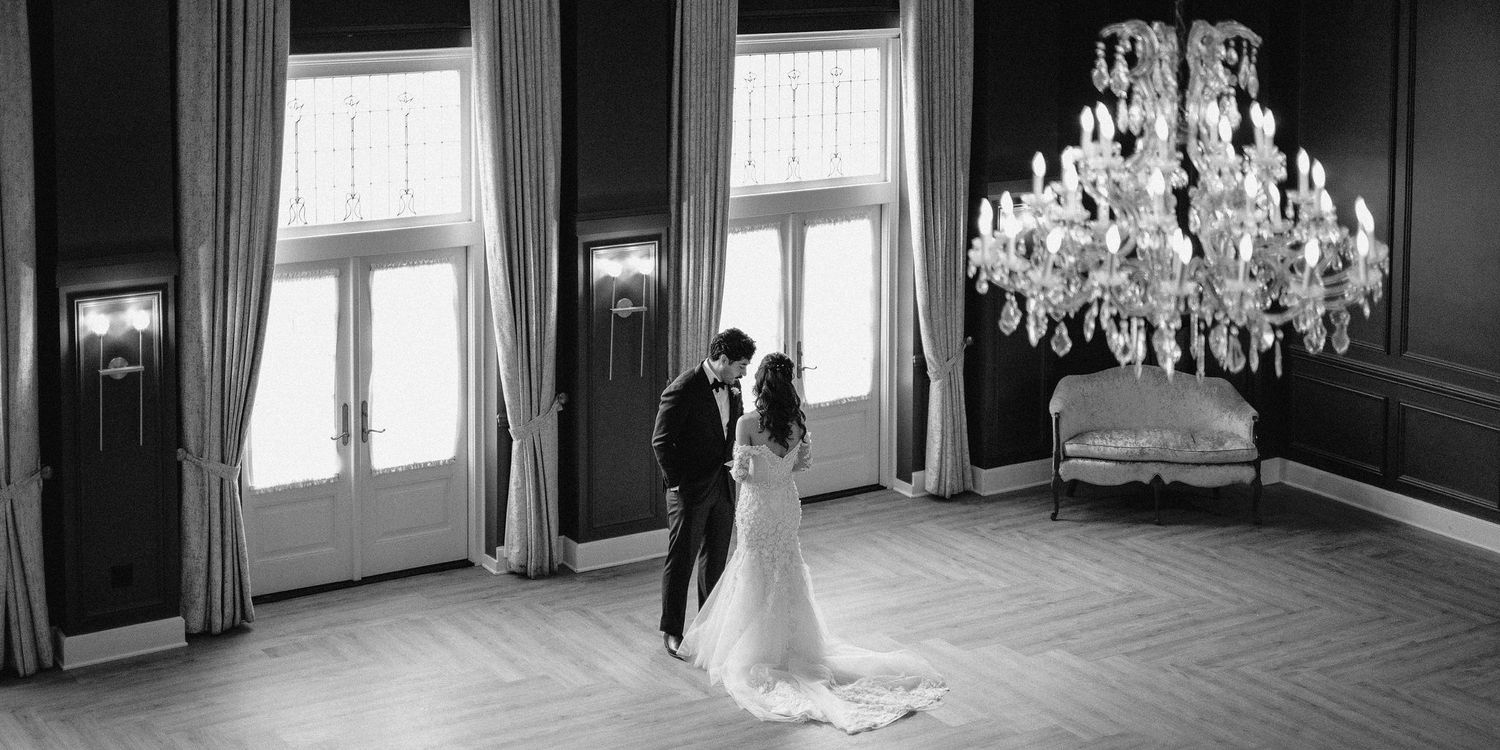 A romantic black and white photograph of a couple in formal attire standing by tall windows beneath an ornate crystal chandelier.