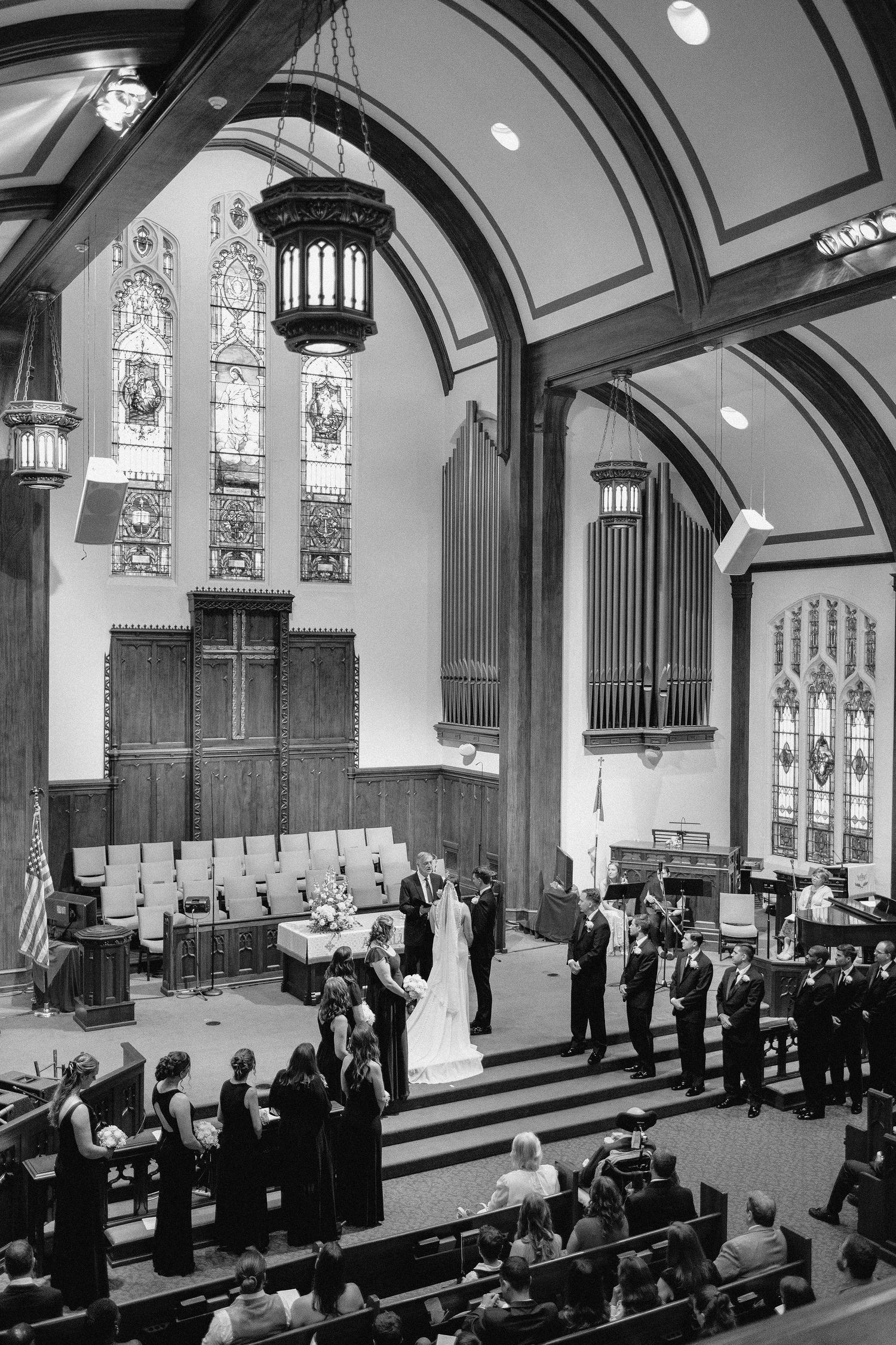 A black and white photograph of a traditional church wedding ceremony with stained glass windows and organ pipes.