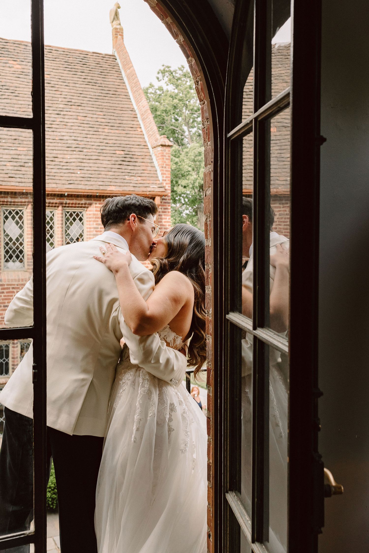 A romantic wedding couple shares a tender kiss in a sunlit church doorway surrounded by dark wooden frames.