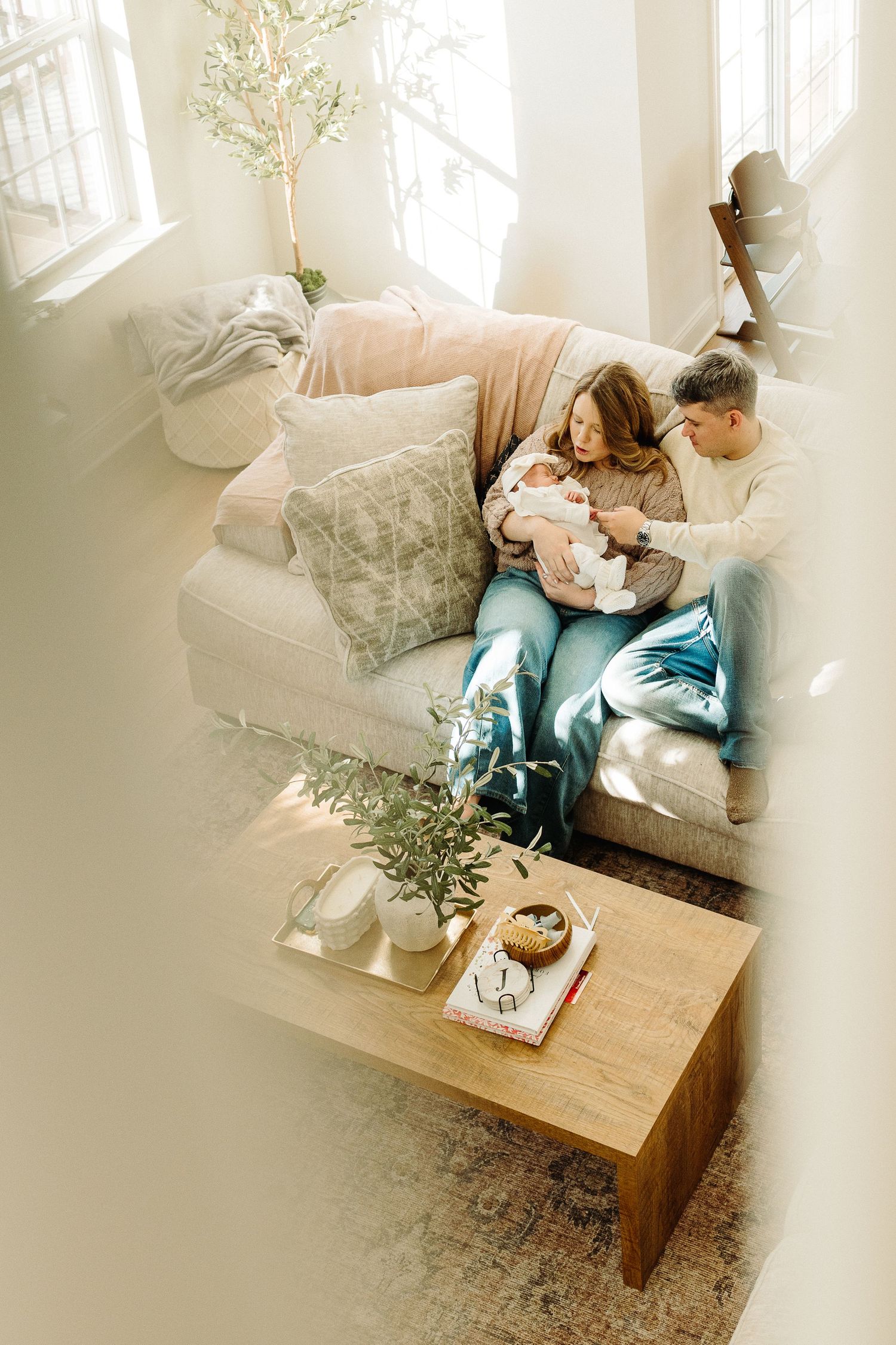 Couple sitting on a cozy sofa cradling their newborn baby in a bright, sunlit living room with neutral decor.