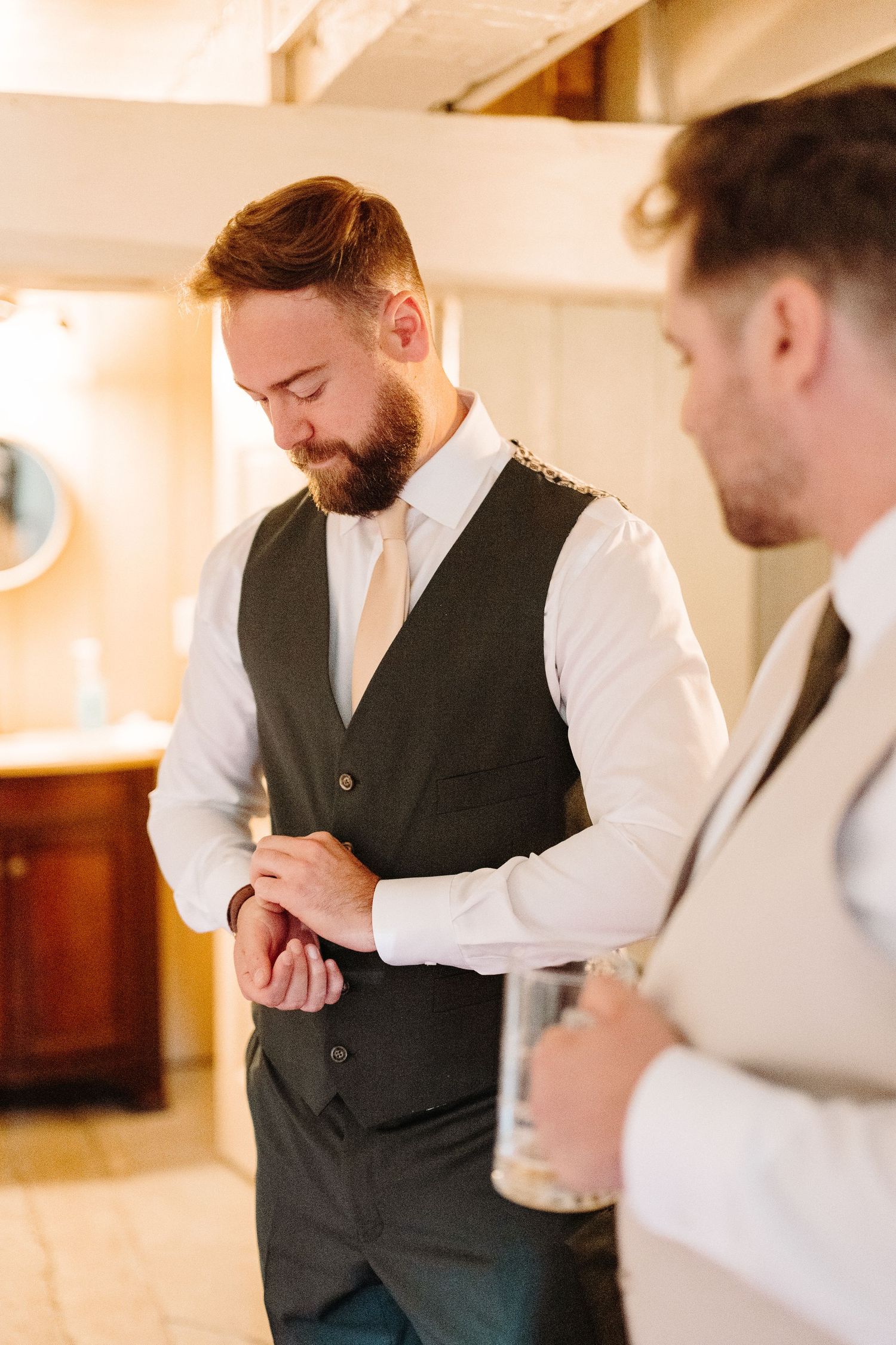 A group of men in formal attire gather together, wearing white shirts and dark vests in a warmly lit indoor setting.