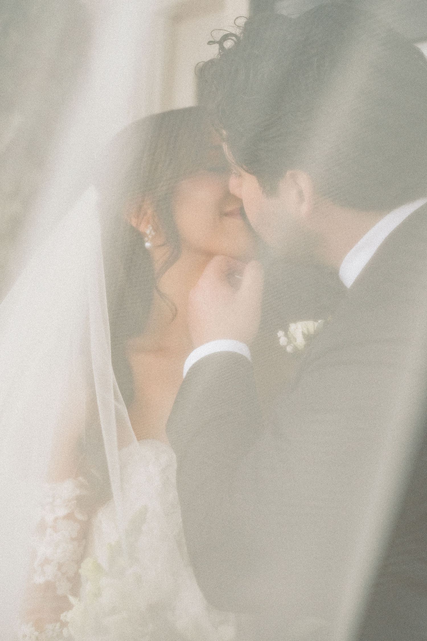 A romantic wedding moment captured through soft focus as the bride and groom share an intimate kiss under a veil.