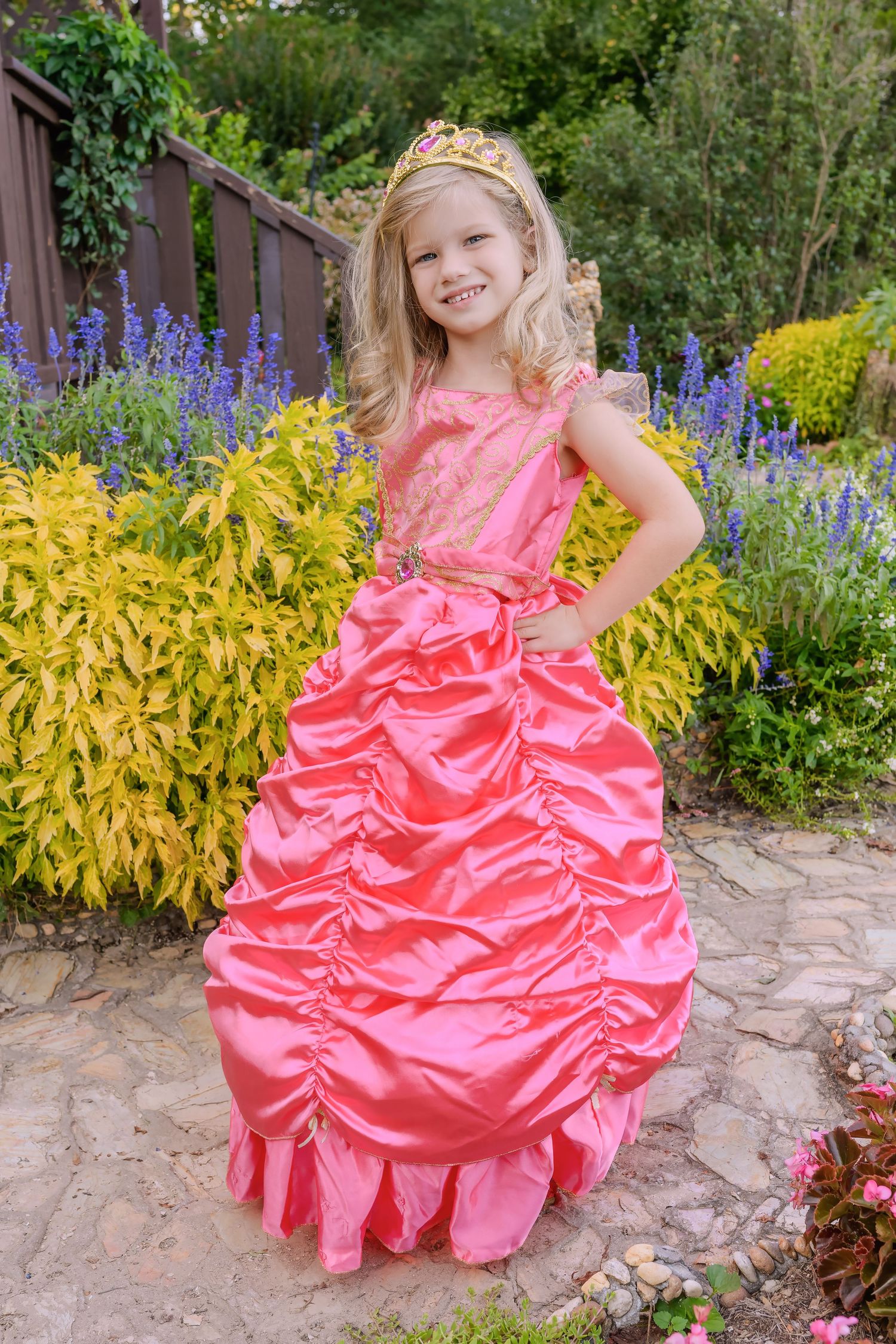 Young blonde girl in pink princess gown and tiara posing in a colorful garden.