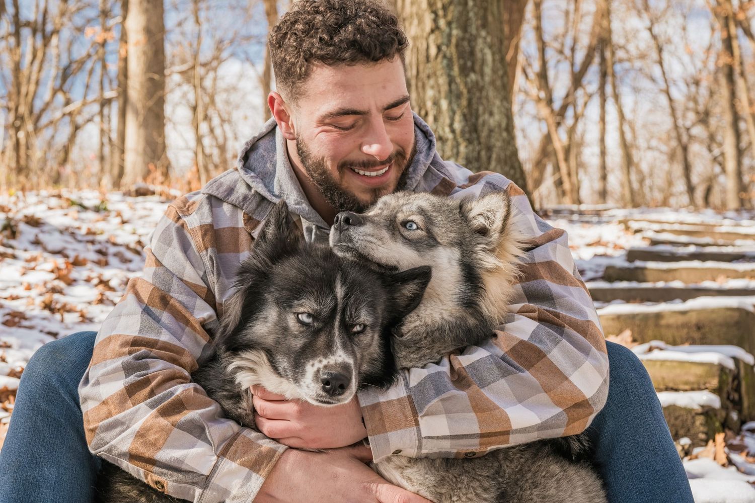 Happy bearded man hugging two Alaskan Malamute dogs in a snowy winter forest setting.