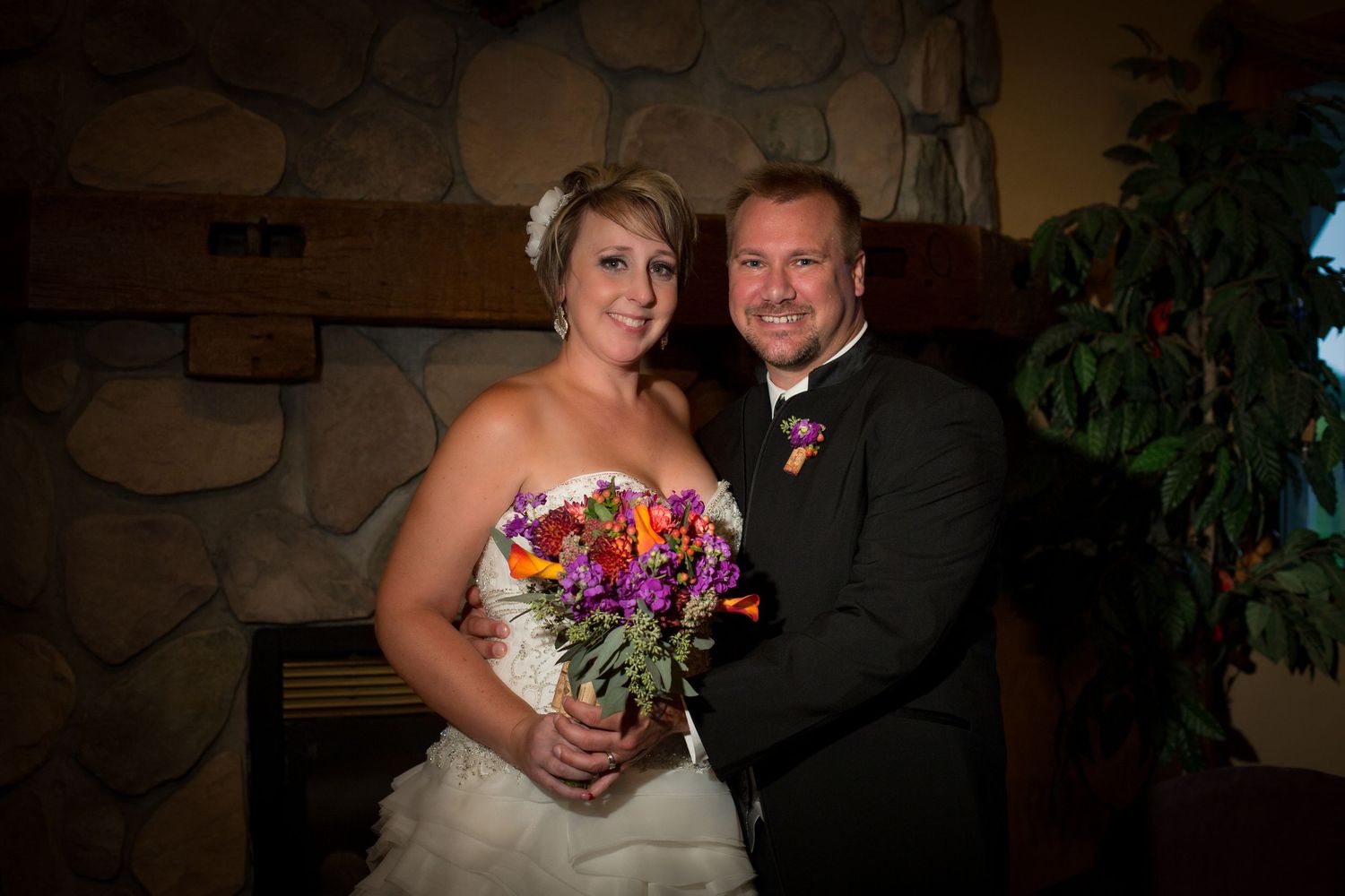 Smiling bride and groom posing indoors by stone fireplace with colorful bouquet.