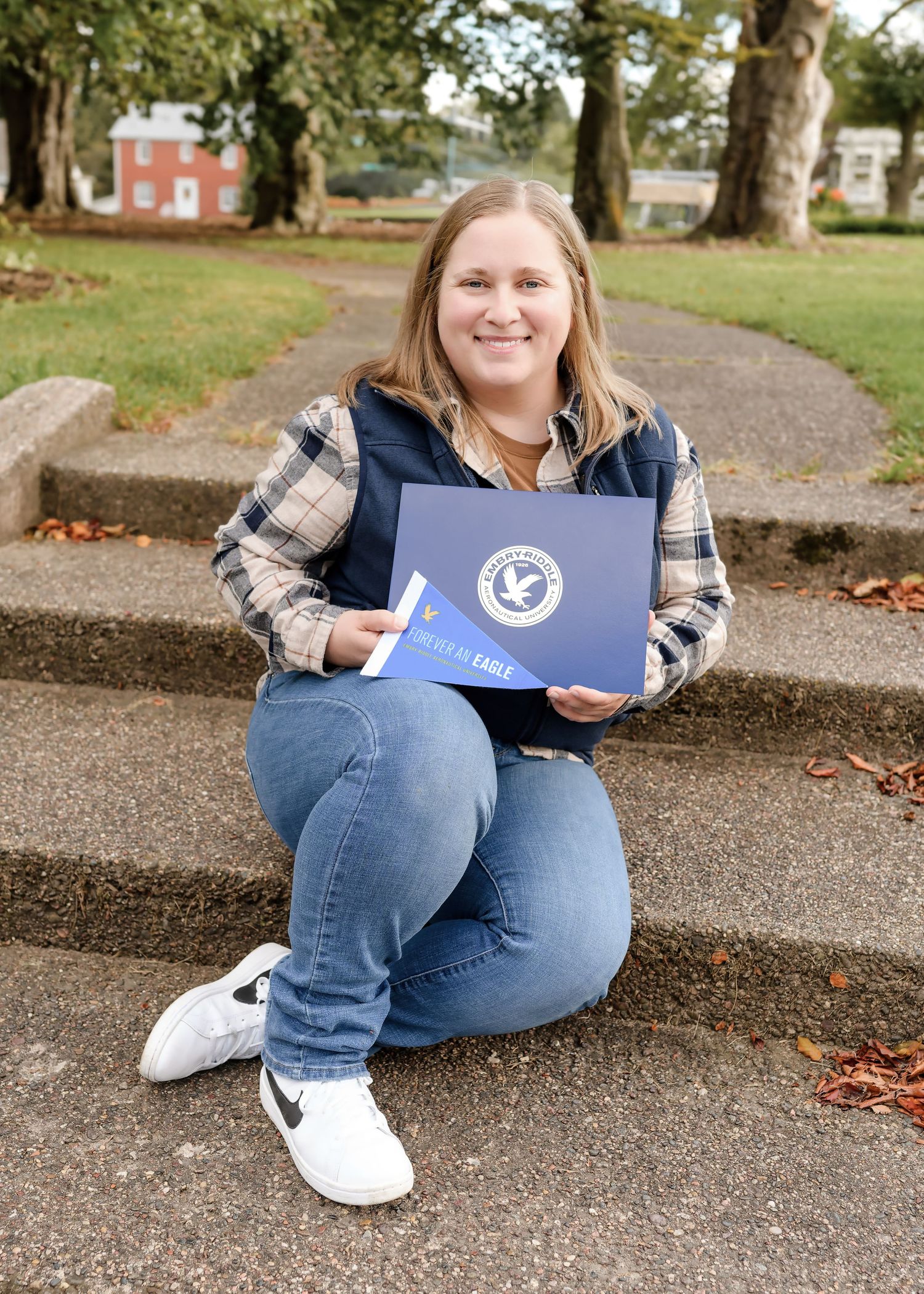 Young woman sitting on steps holding a blue college folder, wearing jeans and a plaid shirt outdoors.