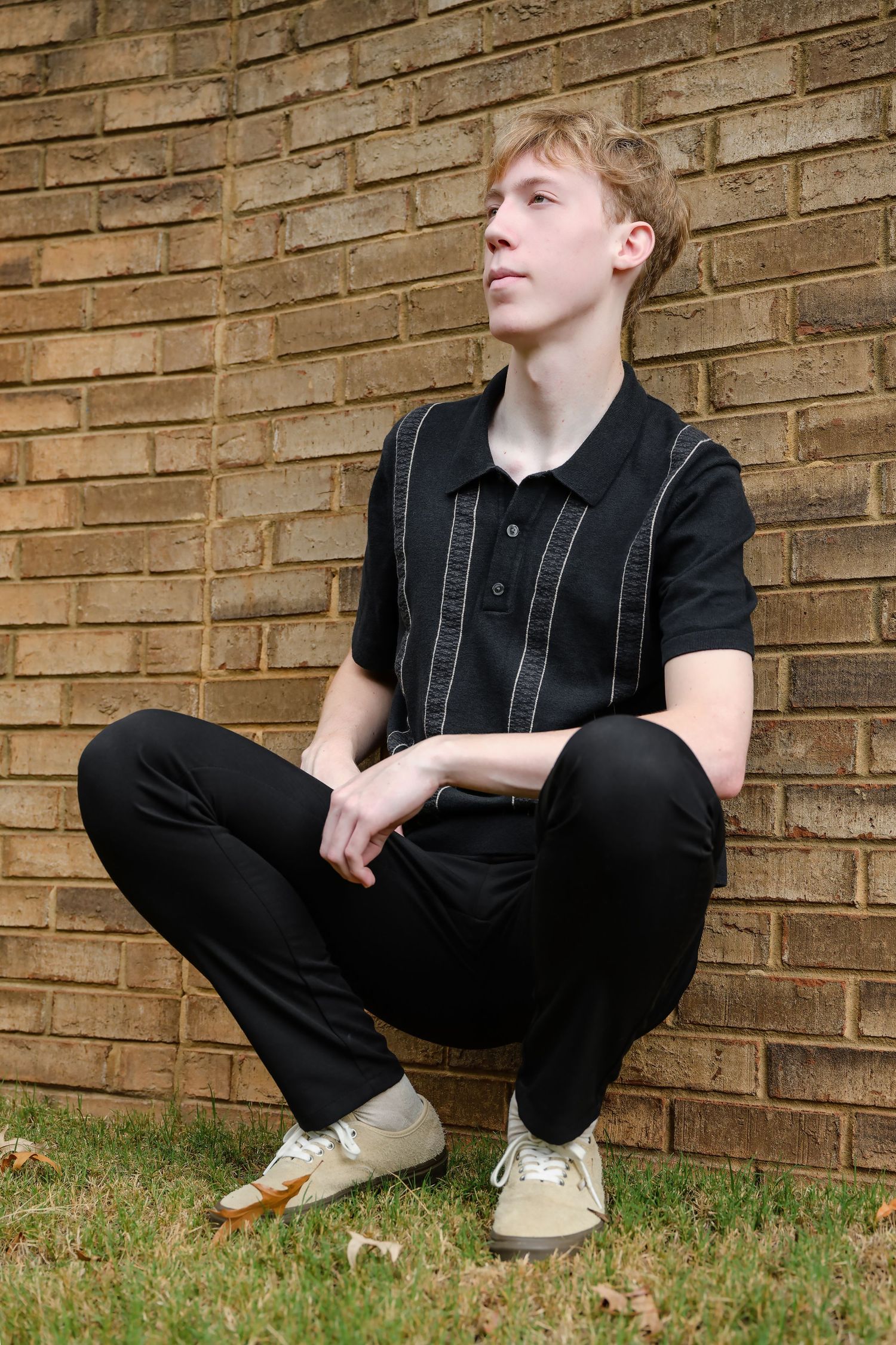 Young blond teen in black polo crouching against a brick wall outdoors.