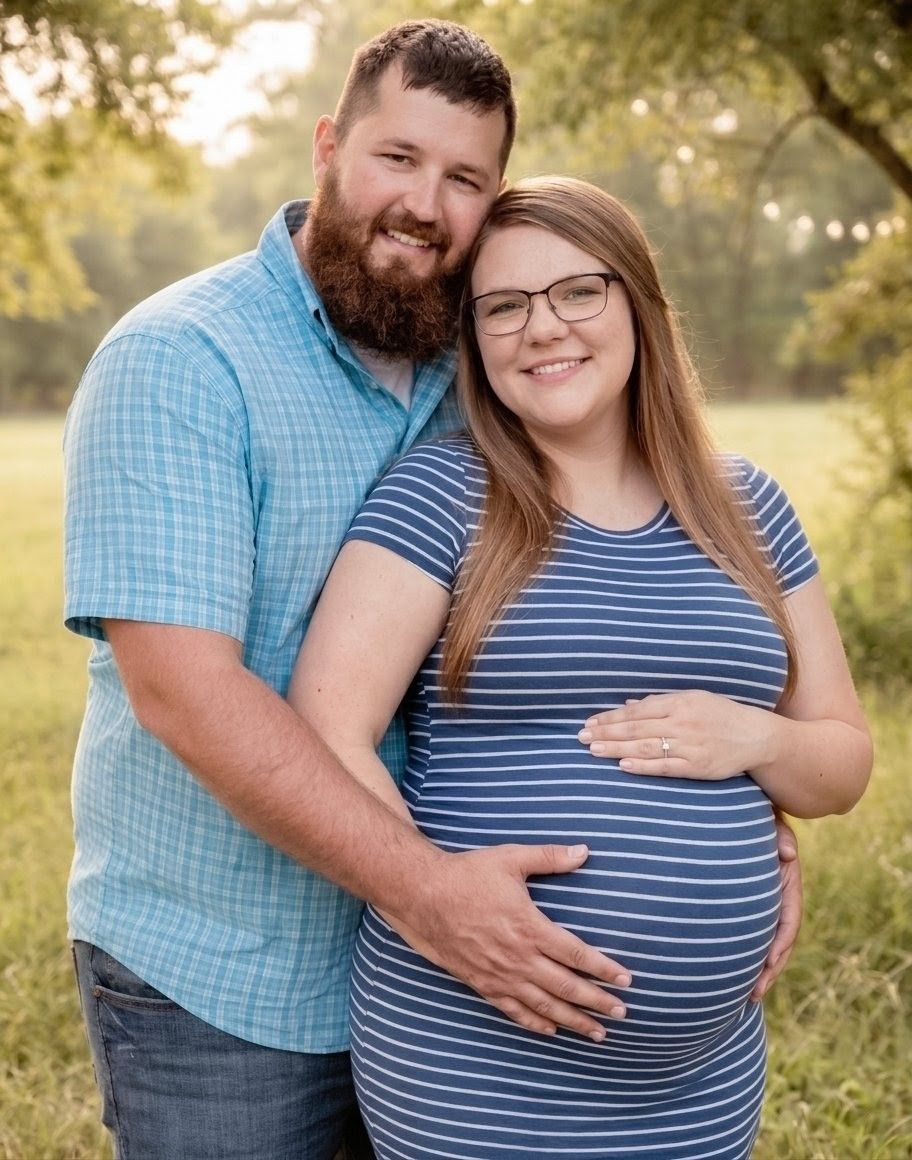 Smiling pregnant couple embracing outdoors in a sunlit green field at golden hour.