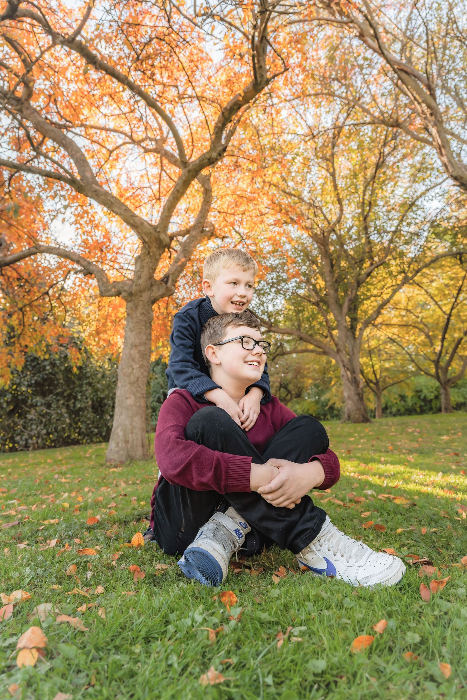Two boys sitting on grass in autumn park, older boy wearing glasses smiling, younger boy hugging him from behind.