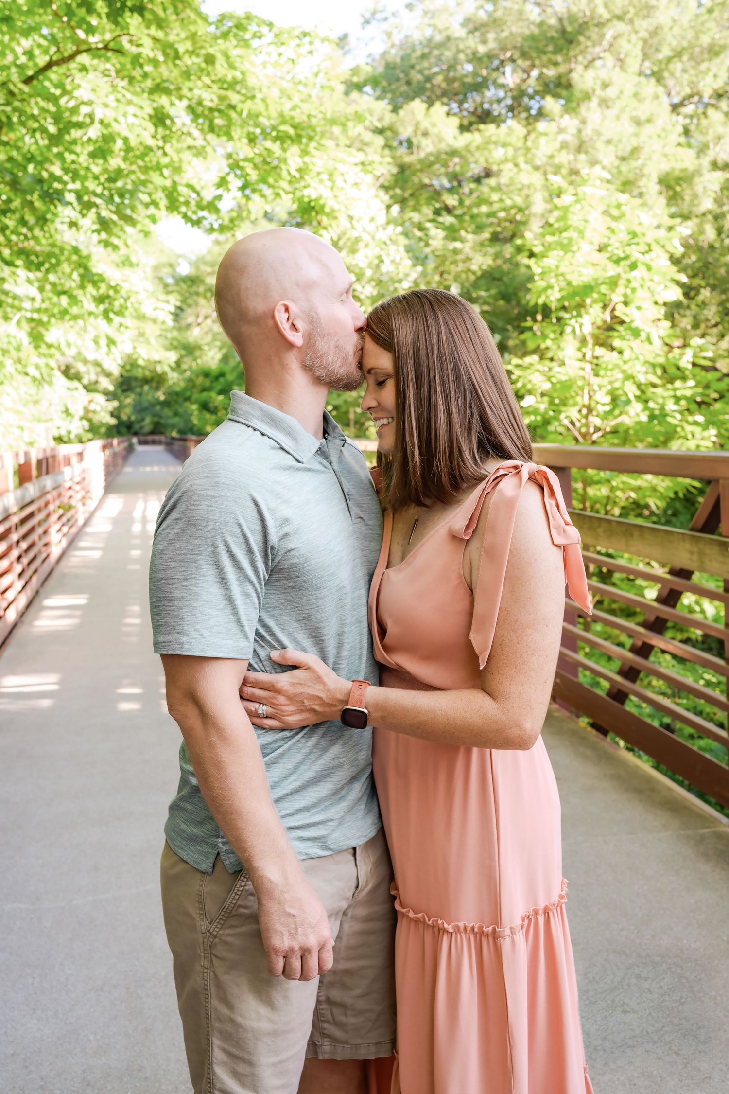 Couple sharing a forehead kiss on a wooden bridge surrounded by lush green trees in summer.