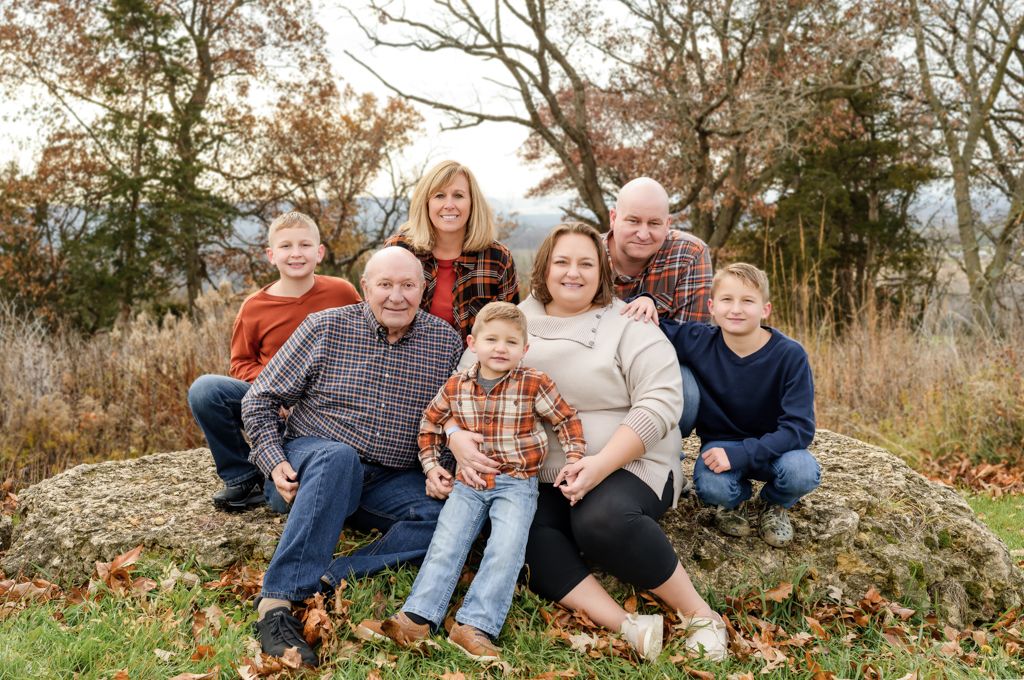 Multi-generational family of seven smiling together outdoors on rocks surrounded by autumn foliage.