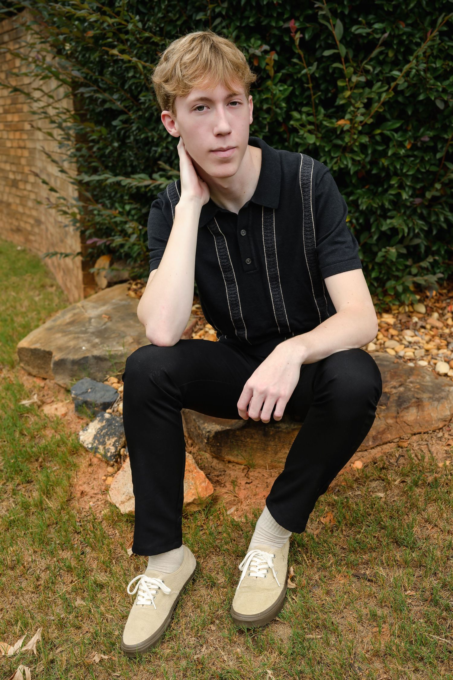 Young blonde teen boy in black outfit sitting on rock outdoors with hand on cheek.