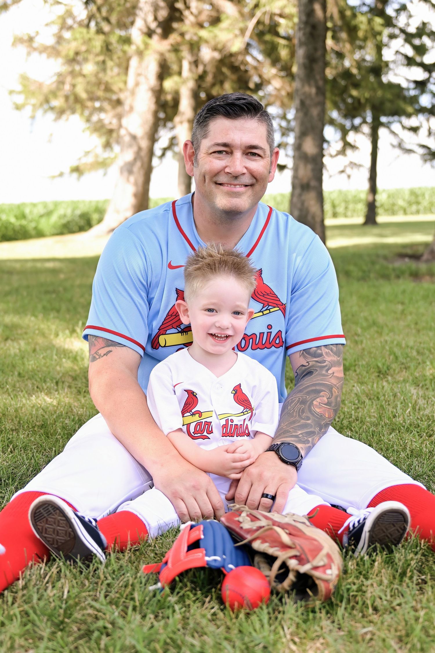 Man and young boy in matching St. Louis Cardinals baseball uniforms sitting together on grass with baseball gear.