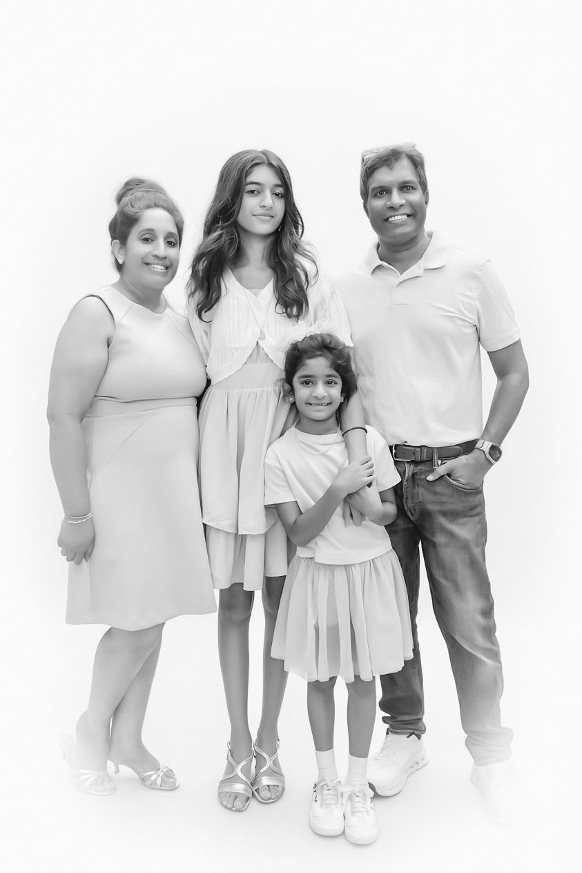 Black and white family portrait of four smiling together, parents with two daughters posing against white background.