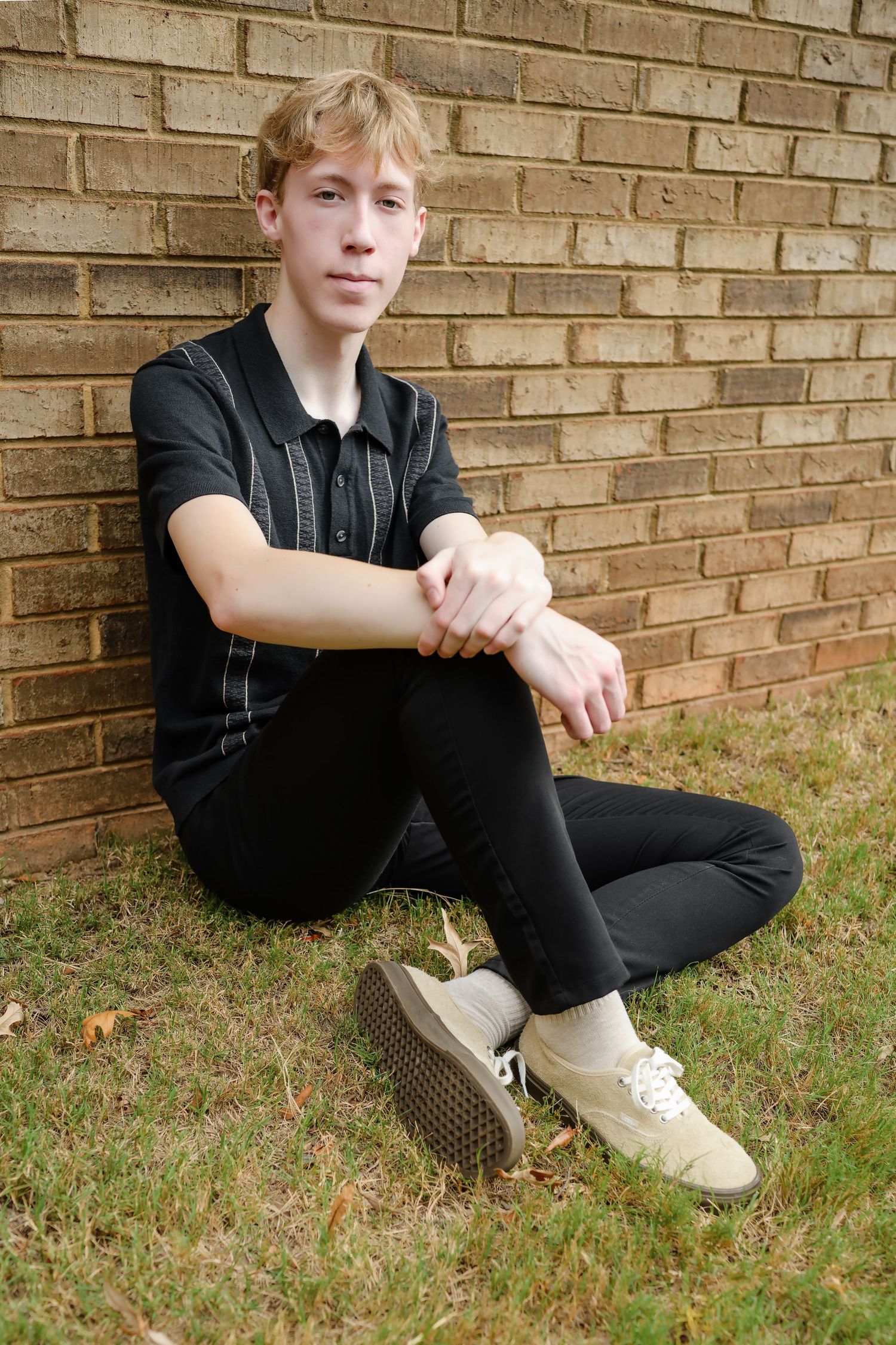 Teenage boy in black outfit sitting against a brick wall on green grass.